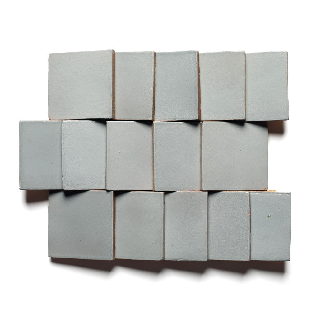 Fourteen Roca 4x4 tiles by Zia Tile, in light gray, are arranged in a staggered, brick-like pattern on a white background. The handmade Cotto-style tiles form two uneven rows with some edges slightly protruding.