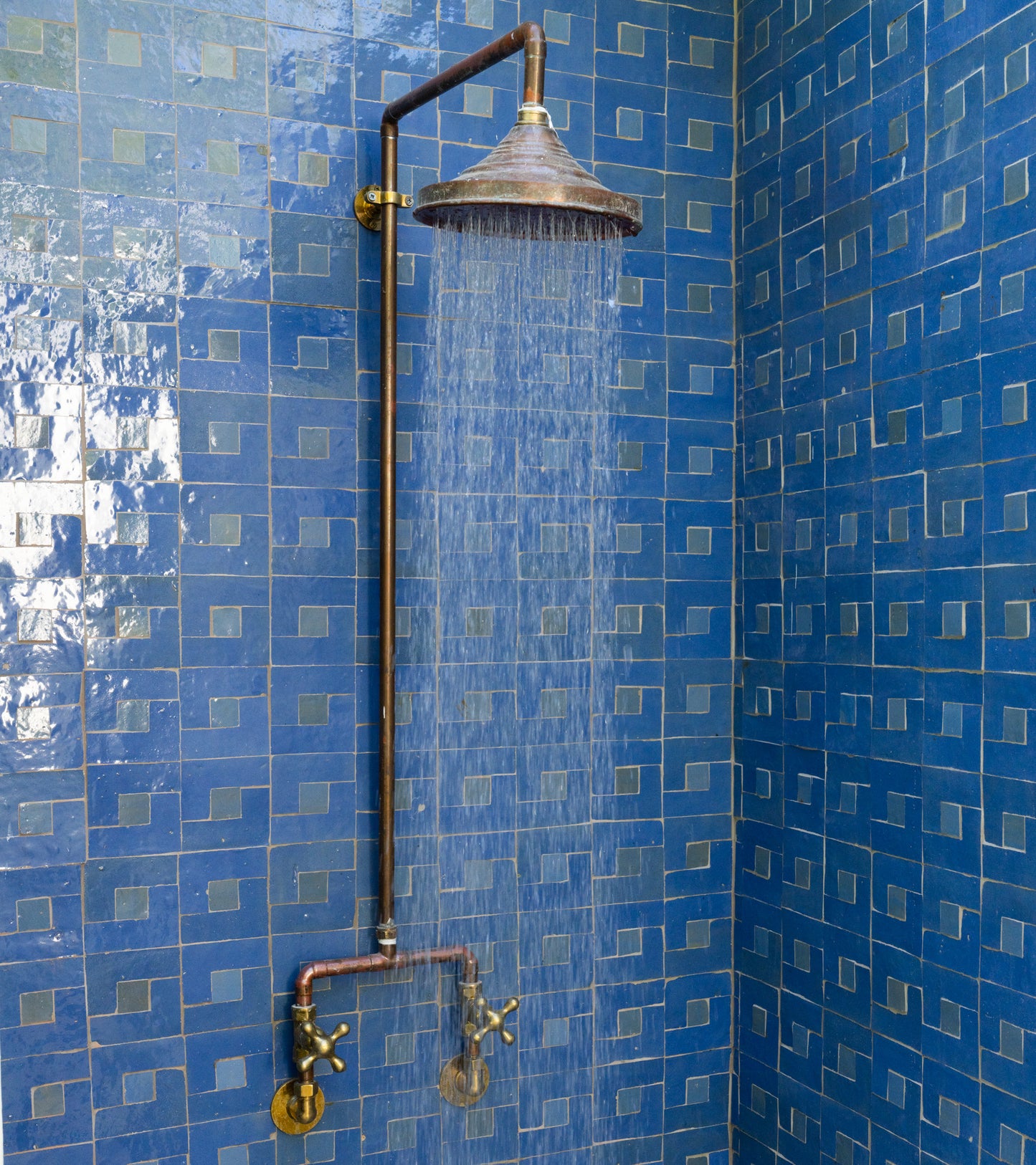 A shower with a large, round metal showerhead and exposed copper pipes runs against a wall clad in Rubric 7 blue mosaic zellige tiles by Zia Tile, featuring a striking geometric pattern.