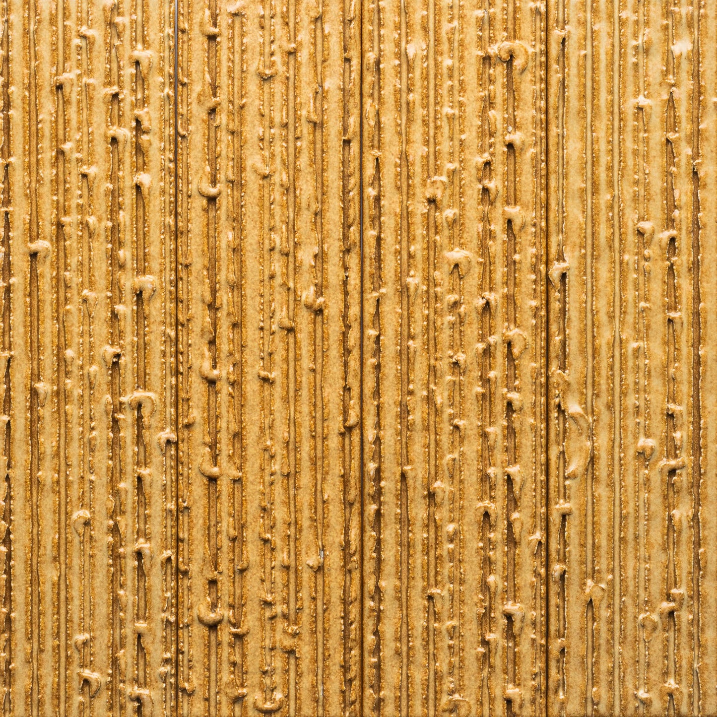 A close-up of Toast + Furrowed by Zia Tile, showing a golden-brown textured surface with vertical, uneven lines and raised bumps, creating a sand-like or heavily frosted appearance.