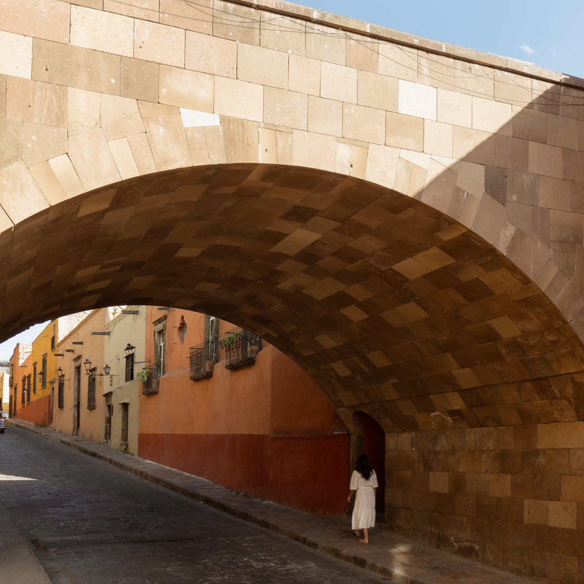 A person in white walks under a large archway made of Sierra 12x24 by Zia Tile on a quiet, sunlit street lined with colorful buildings.