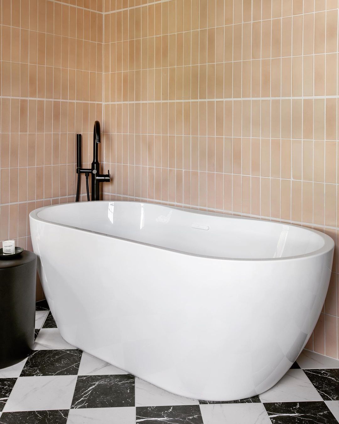 A modern bathroom featuring a white freestanding tub, Jaipur Pink 2x8 tiles on the walls, black fixtures, a small round black side table, and black-and-white checkered floor tiles.
