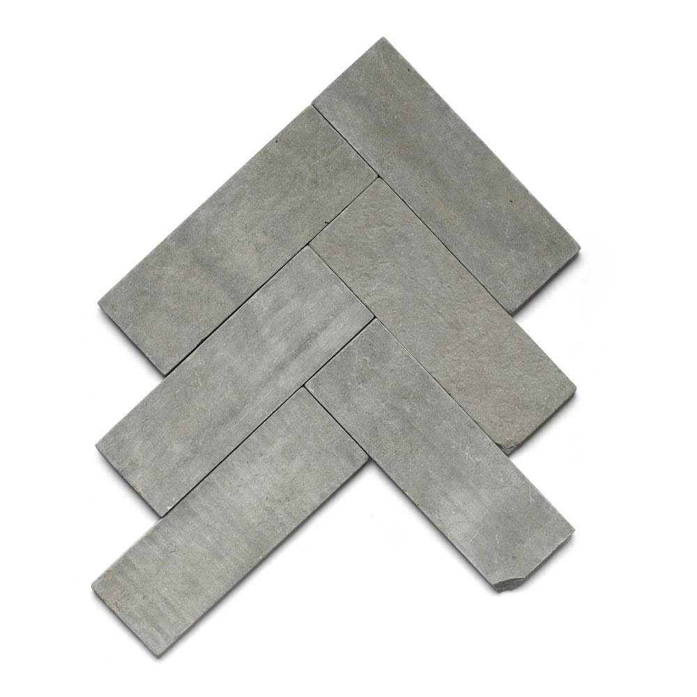 Five Basilica 4x12 + Honed rectangular gray stone tiles, inspired by Moroccan Limestone, are arranged in a herringbone pattern on a white background. The large format tiles have a smooth matte finish with subtle color variation and natural texture.