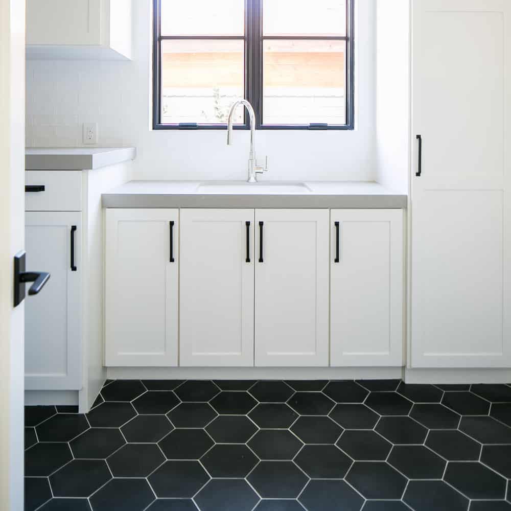 A modern kitchen showcases white cabinets with black handles, a light gray countertop, stainless steel faucet, and bold Black Hex tile flooring. Two windows above the sink bathe the space in natural light.