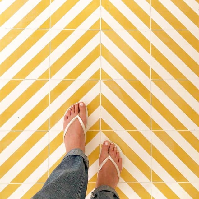 A person wearing white flip-flops and blue jeans stands on an Ocotillo Cadmium 8x8 yellow and white diagonal striped modern tile floor.