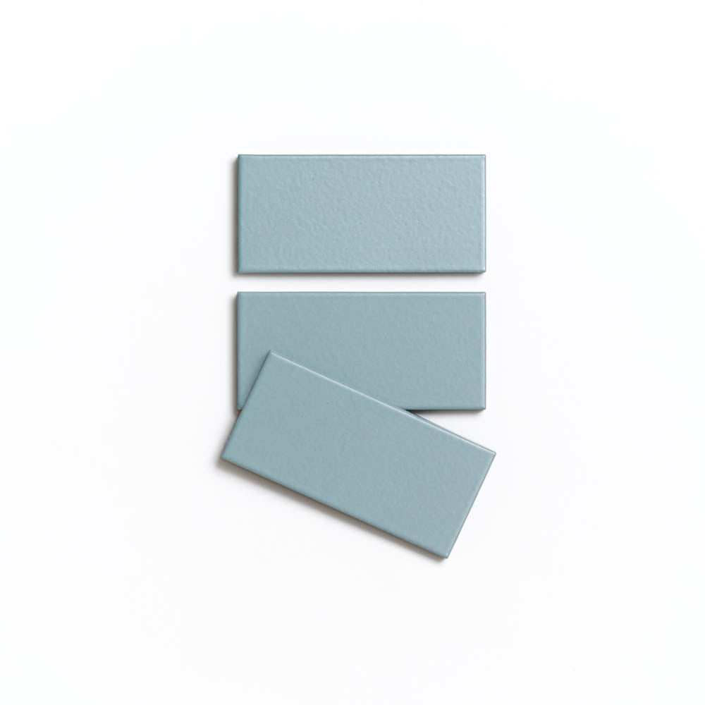 Three Moonlight 2x4 tiles by Zia Tile, in light blue matte ceramic, are arranged on a white background: two stacked vertically and one placed diagonally across the lower tile.