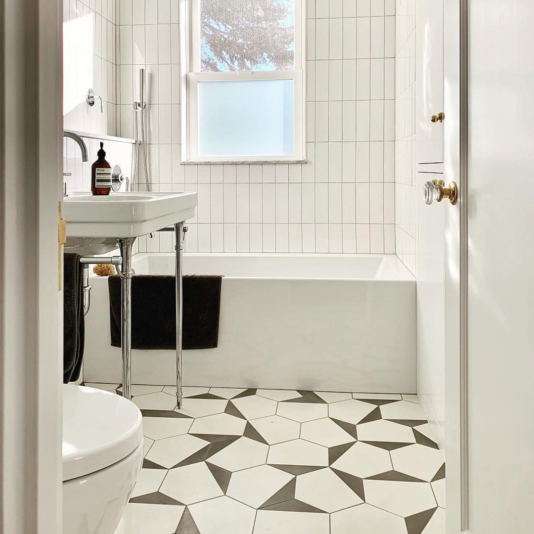 Bright bathroom with white tiled walls, white bathtub, sink, and toilet. The floor features Oslo Hex black and white geometric cement tiles. A frosted window lets in light, and a brown towel with a soap bottle sits by the sink.
