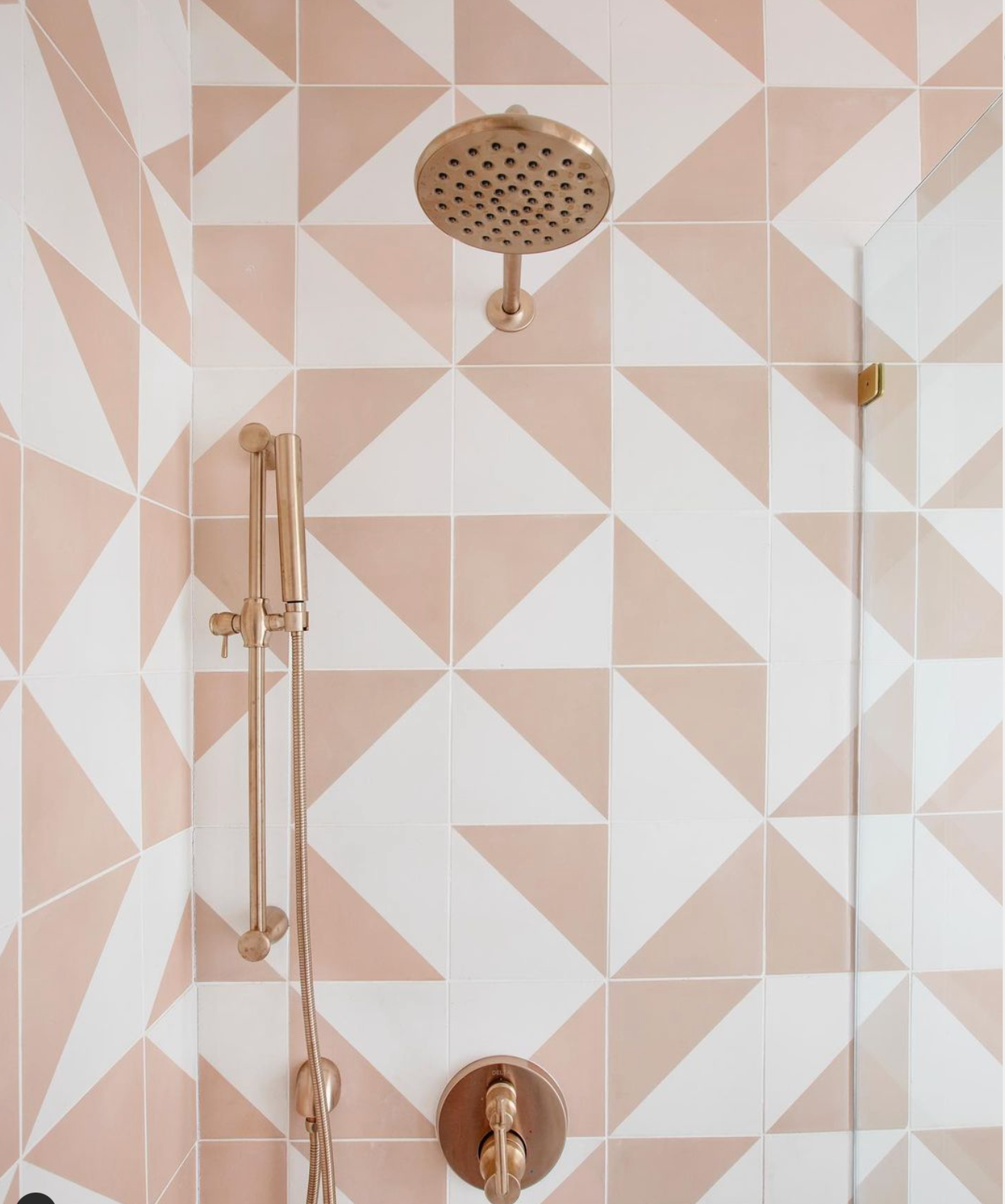 Delta Jaipur Pink 8x8 tiles in geometric pink and white create a stylish shower, paired with modern brass-colored fixtures—a rainfall showerhead, handheld shower, and control knob. A partial glass door appears on the right.