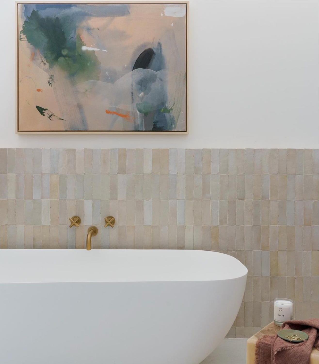 A modern bathroom features a white tub, brass fixtures, Casablanca 2x6 beige Moroccan-inspired tiles by Zia Tile, an abstract painting above, and a pink towel, white candle, and soap on a wooden stool beside the tub.