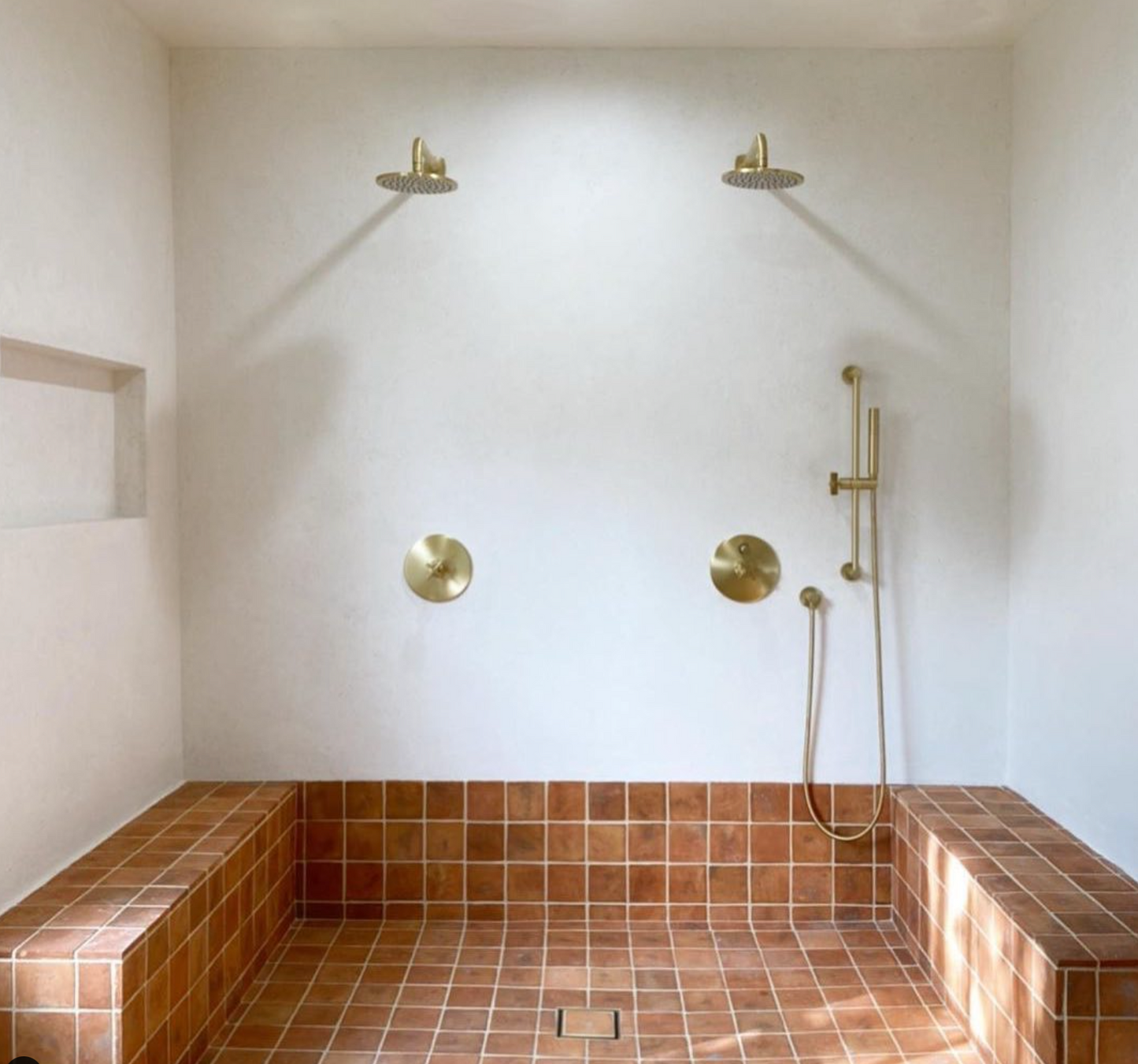 A minimalist shower features dual brass rain showerheads, dual controls, a handheld above a bench clad in Zia Tiles 4x4 Square + Fired Earth handmade tiles, and white walls with a recessed shelf on the left.