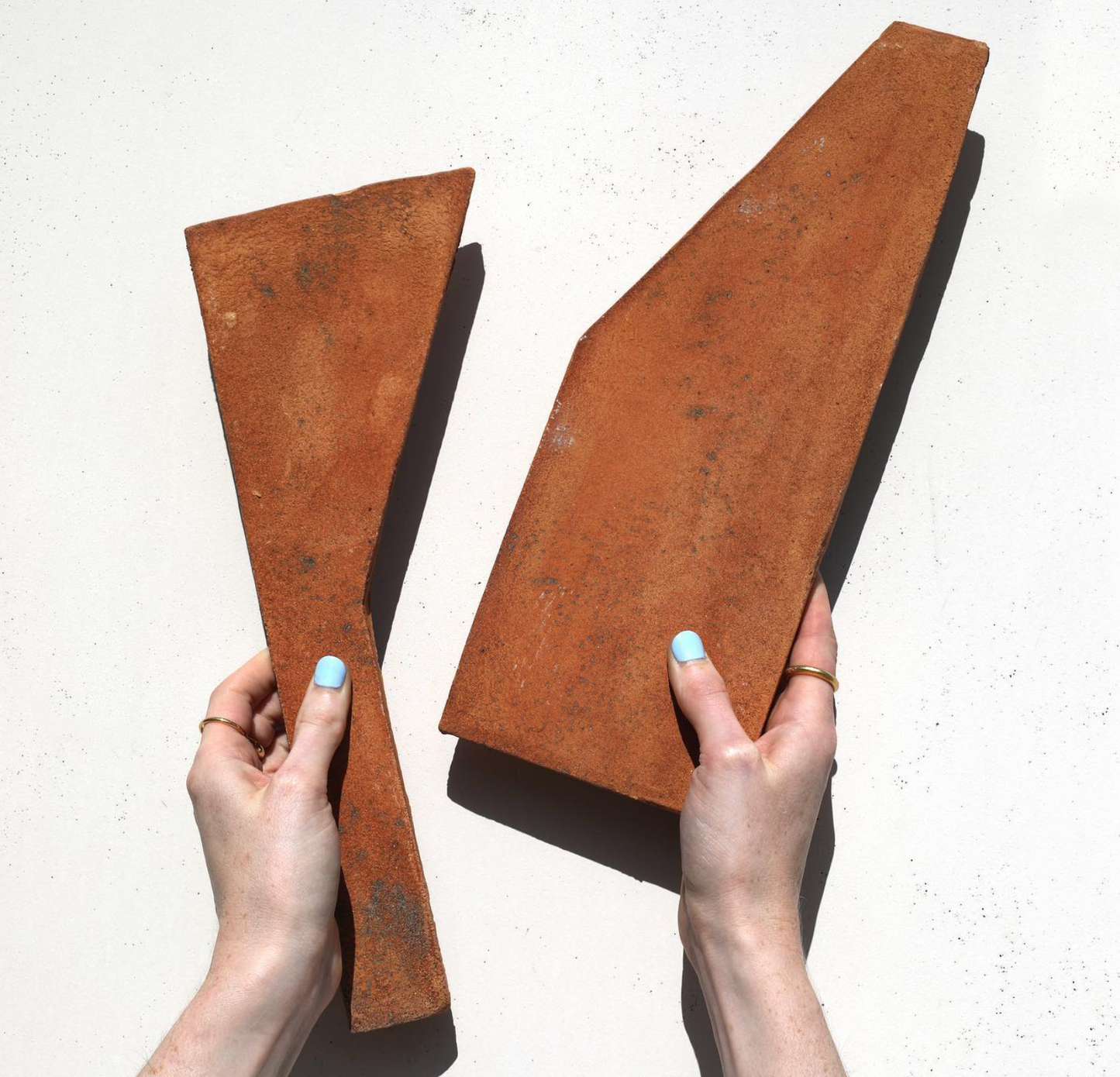 Two hands with light blue nail polish hold two angular, uniquely-shaped Zocalo + Fired Earth bricks by Zia Tile against a white background. Their varying size and artisan finish showcase the handcrafted beauty of ceramic arts.