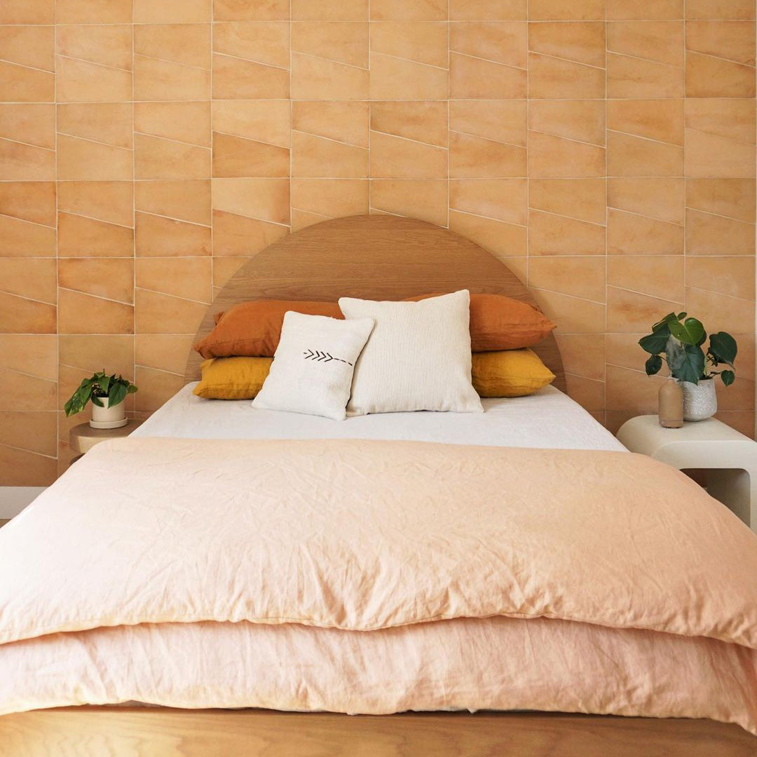 A neatly made bed with peach bedding and a wooden headboard stands against a tan Zia Tile Toltec + Adobe geometric tiled wall. Small plants rest on white bedside tables on either side of the bed.