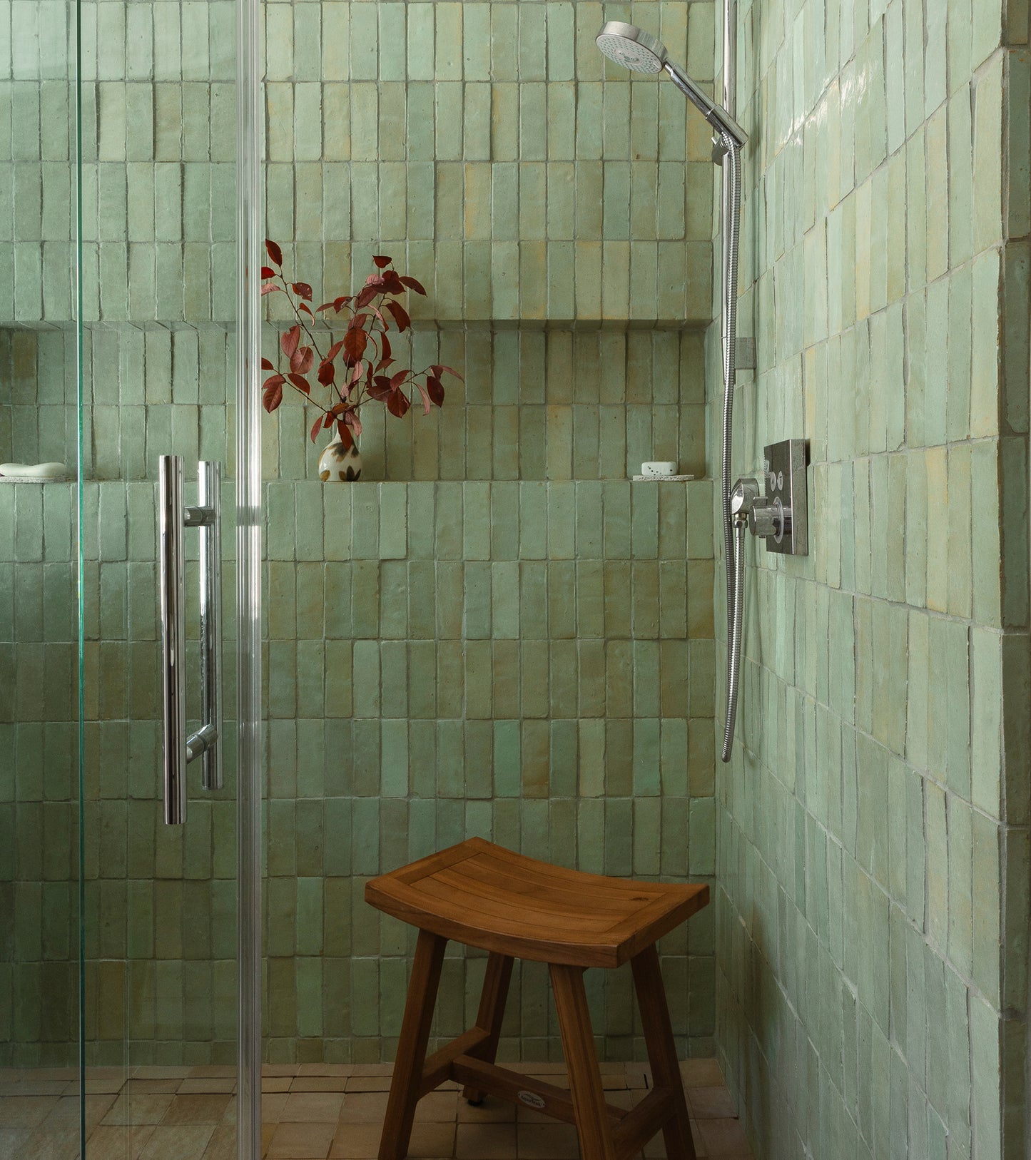 A shower featuring Absinthe 2x6 tiles by Zia Tile, a glass door, wooden stool, built-in shelf with a white vase of red leaves, and modern silver fixtures brings Moroccan-inspired charm to the space.