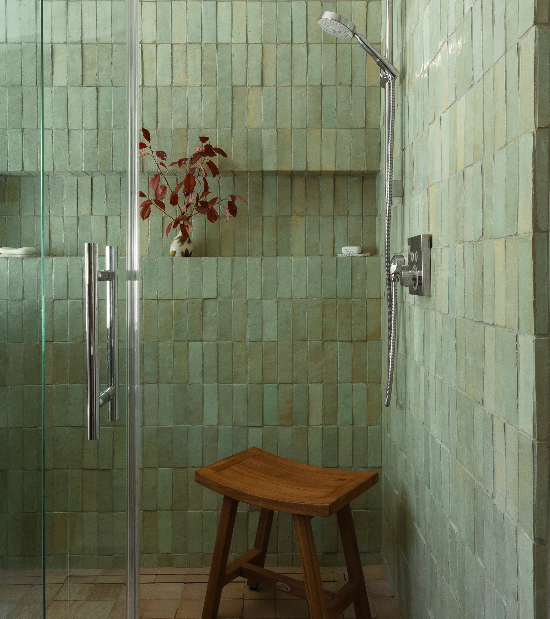 A shower featuring Absinthe 2x6 tiles by Zia Tile, a glass door, wooden stool, built-in shelf with a white vase of red leaves, and modern silver fixtures brings Moroccan-inspired charm to the space.