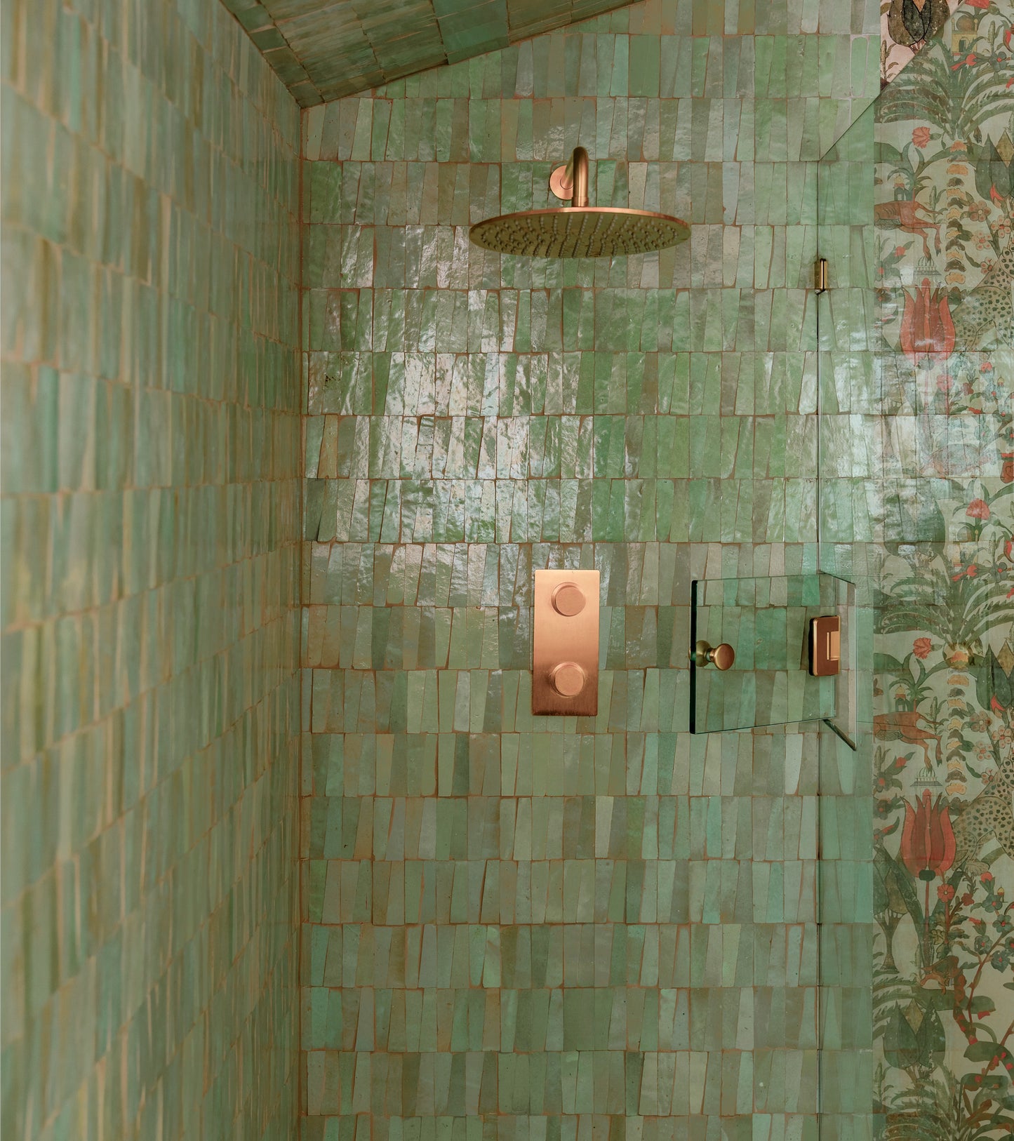 A shower featuring Zia Tile's Absinthe Trapezoid green handmade tiles in a rectangular pattern, with gold fixtures and showerhead, plus a glimpse of floral-patterned wallpaper on the right wall.