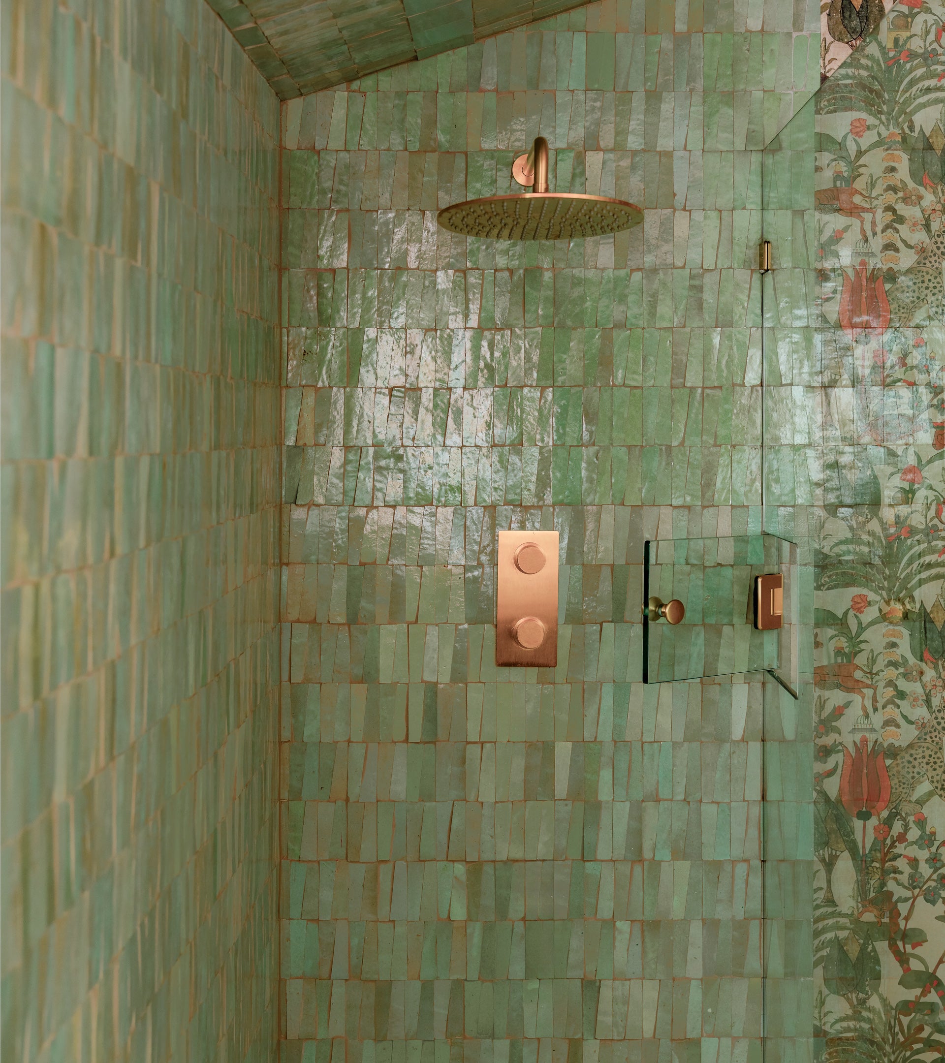 A shower featuring Zia Tile's Absinthe Trapezoid green handmade tiles in a rectangular pattern, with gold fixtures and showerhead, plus a glimpse of floral-patterned wallpaper on the right wall.