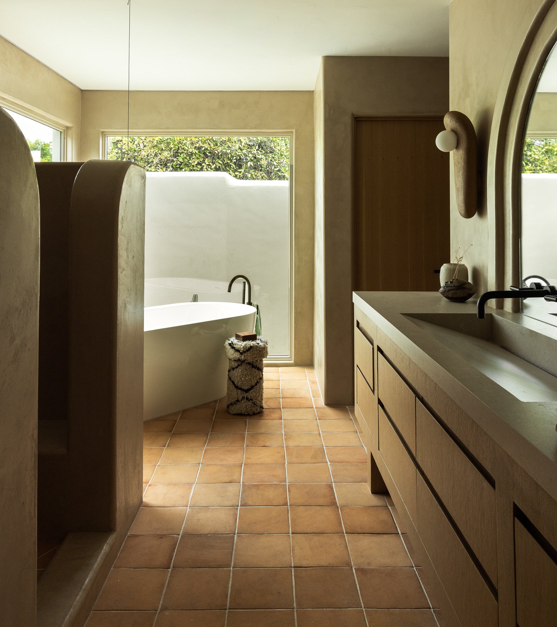 A minimalist bathroom featuring Zia Tile’s 8x8 Square + Adobe terra cotta floor, a freestanding tub by large windows, a curved shower partition, wooden double-sink vanity, and soft natural light inspired by San Miguel de Allende.