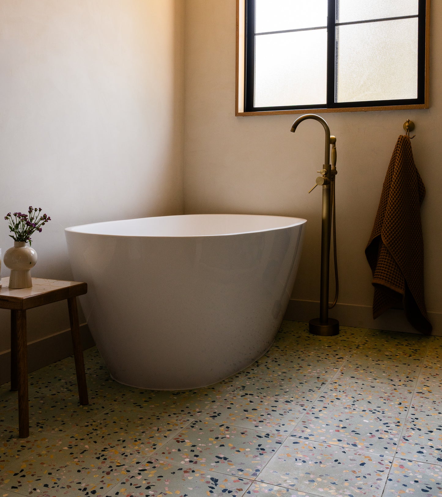 A modern bathroom with a white freestanding tub, tall gold faucet, small wooden stool with a vase, towel on the wall, frosted window above, and Zia Tiles Savoye 12x12 terrazzo cement tiles on the floor.