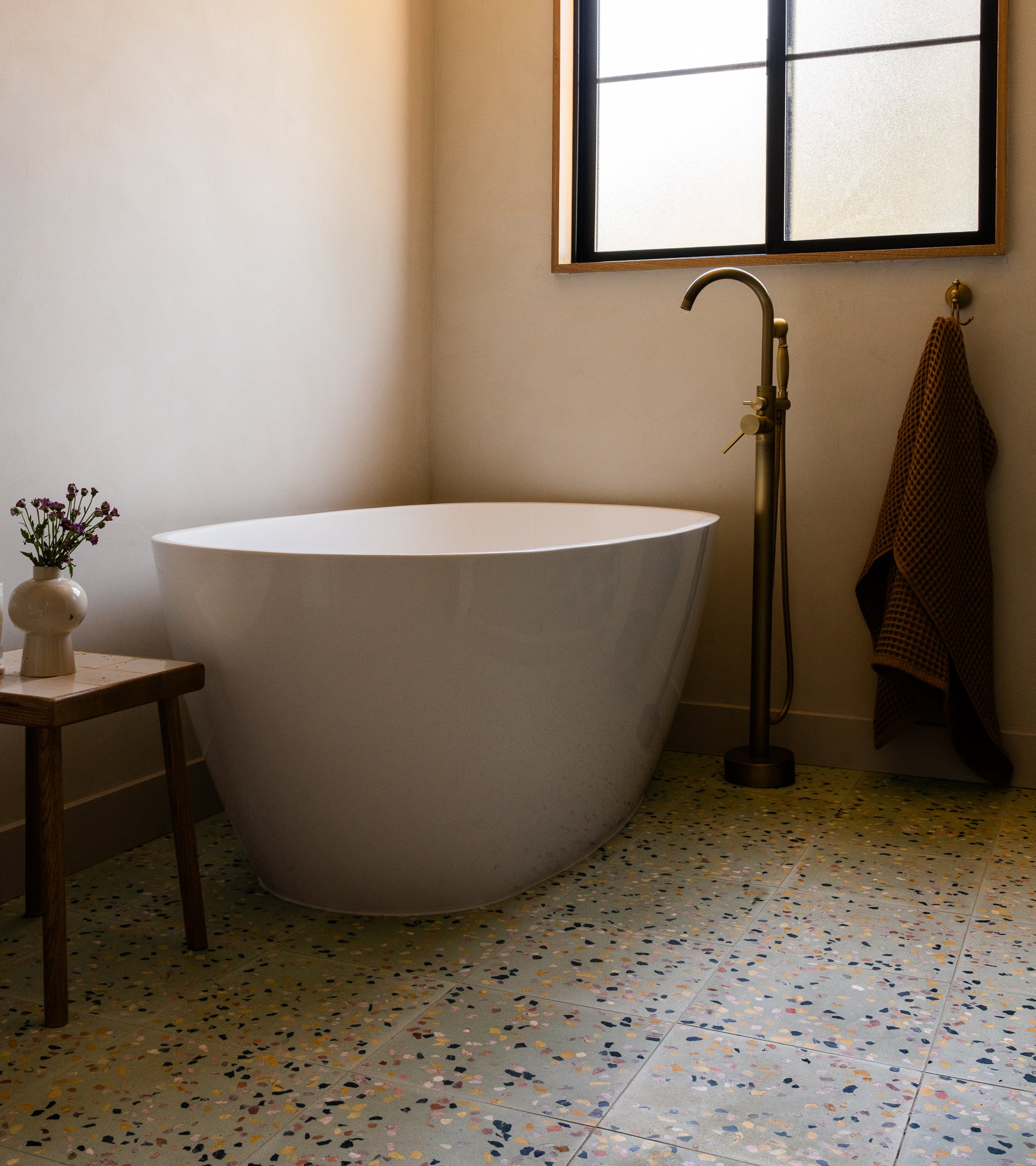 A modern bathroom with a white freestanding tub, tall gold faucet, small wooden stool with a vase, towel on the wall, frosted window above, and Zia Tiles Savoye 12x12 terrazzo cement tiles on the floor.