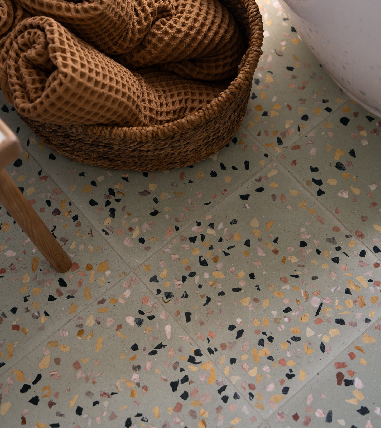 A woven basket with textured brown towels rests on a Savoye 12x12 terrazzo cement tile floor by Zia Tile, featuring multicolored specks, beside the base of a white bathtub.