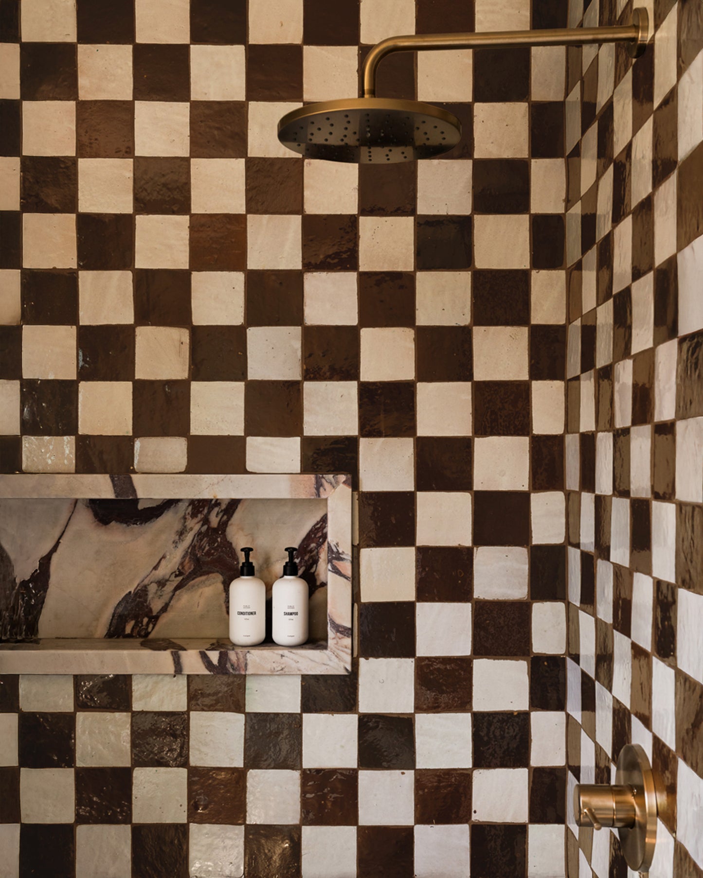 A shower features glossy Burnt Sugar 4x4 tiles in a brown and white checkerboard pattern, a brass showerhead and handle, plus a recessed shelf with two white bottles topped with black pumps. Brand: vendor-unknown.