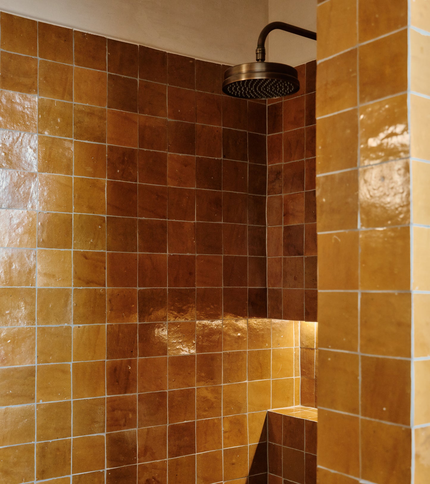 A shower features vendor-unknown’s Amber 4x4 tiles, covering the walls and built-in bench. The glossy brown tiles and round metallic showerhead create a warm, cohesive look inspired by classic Moroccan design.