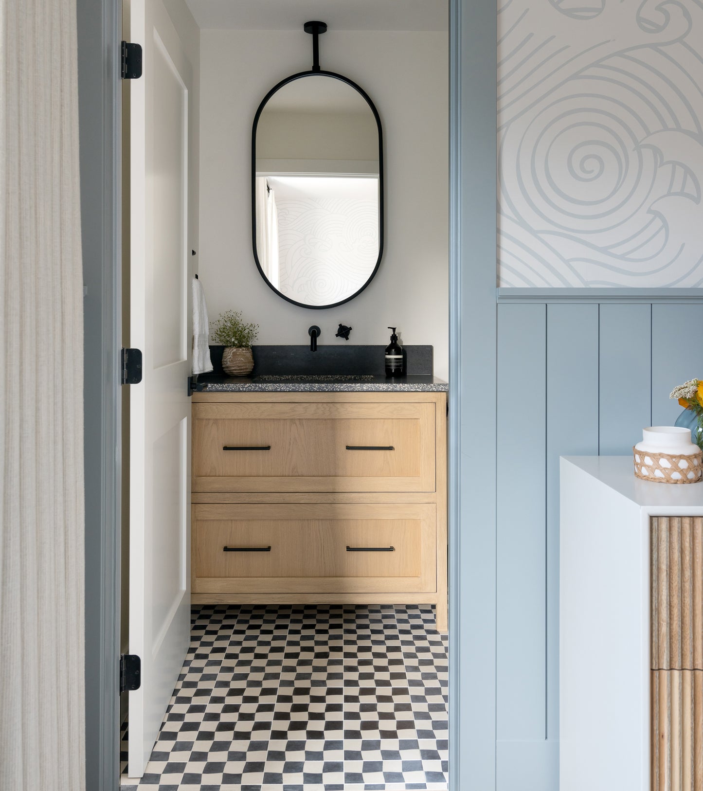 A small bathroom features Zia Tile’s Reality Check Black 4x8 tile on the floor, a light wood vanity with black fixtures and countertop, an oval mirror above, and modern patterned light-colored walls.