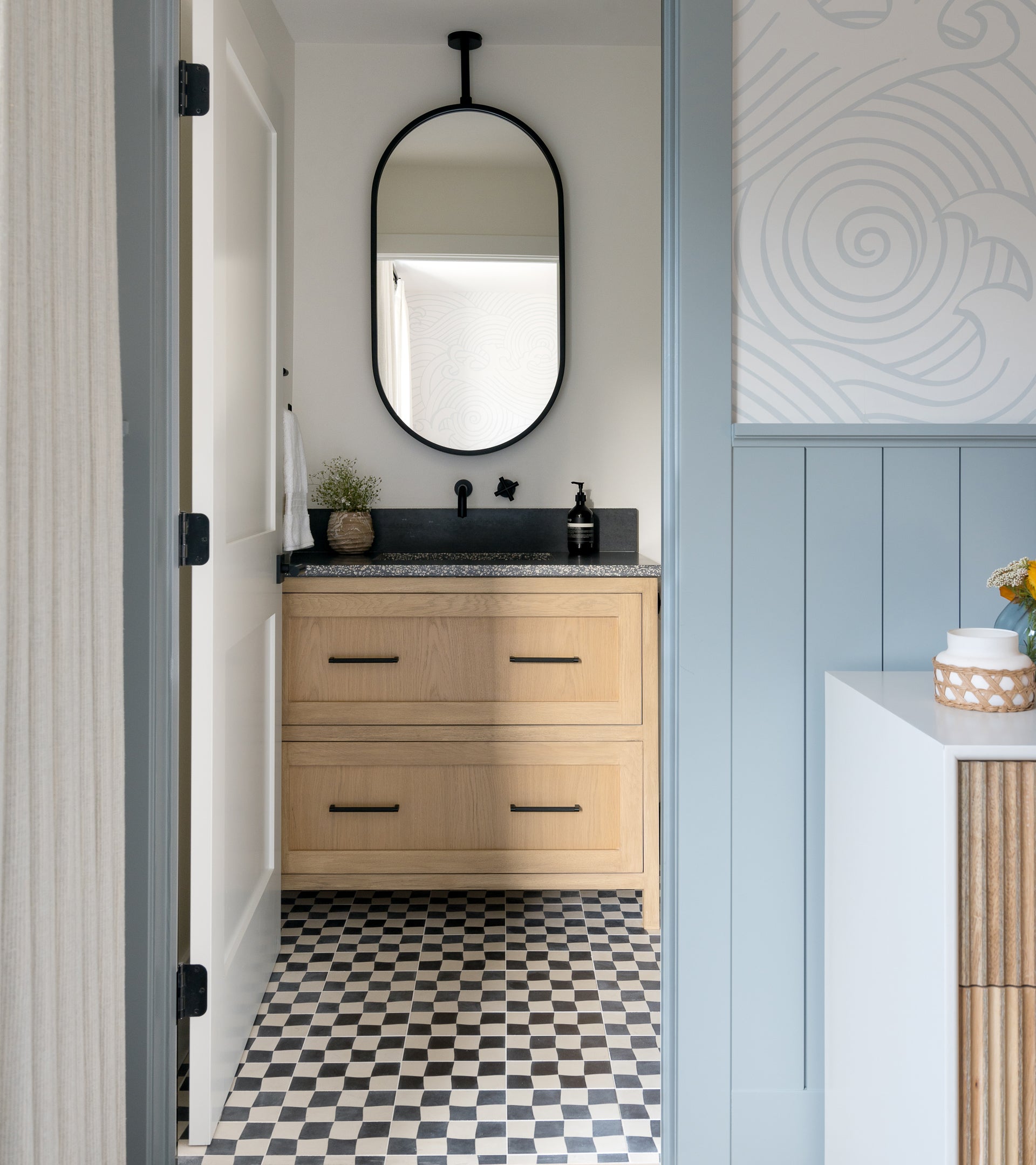 A small bathroom features Zia Tile’s Reality Check Black 4x8 tile on the floor, a light wood vanity with black fixtures and countertop, an oval mirror above, and modern patterned light-colored walls.