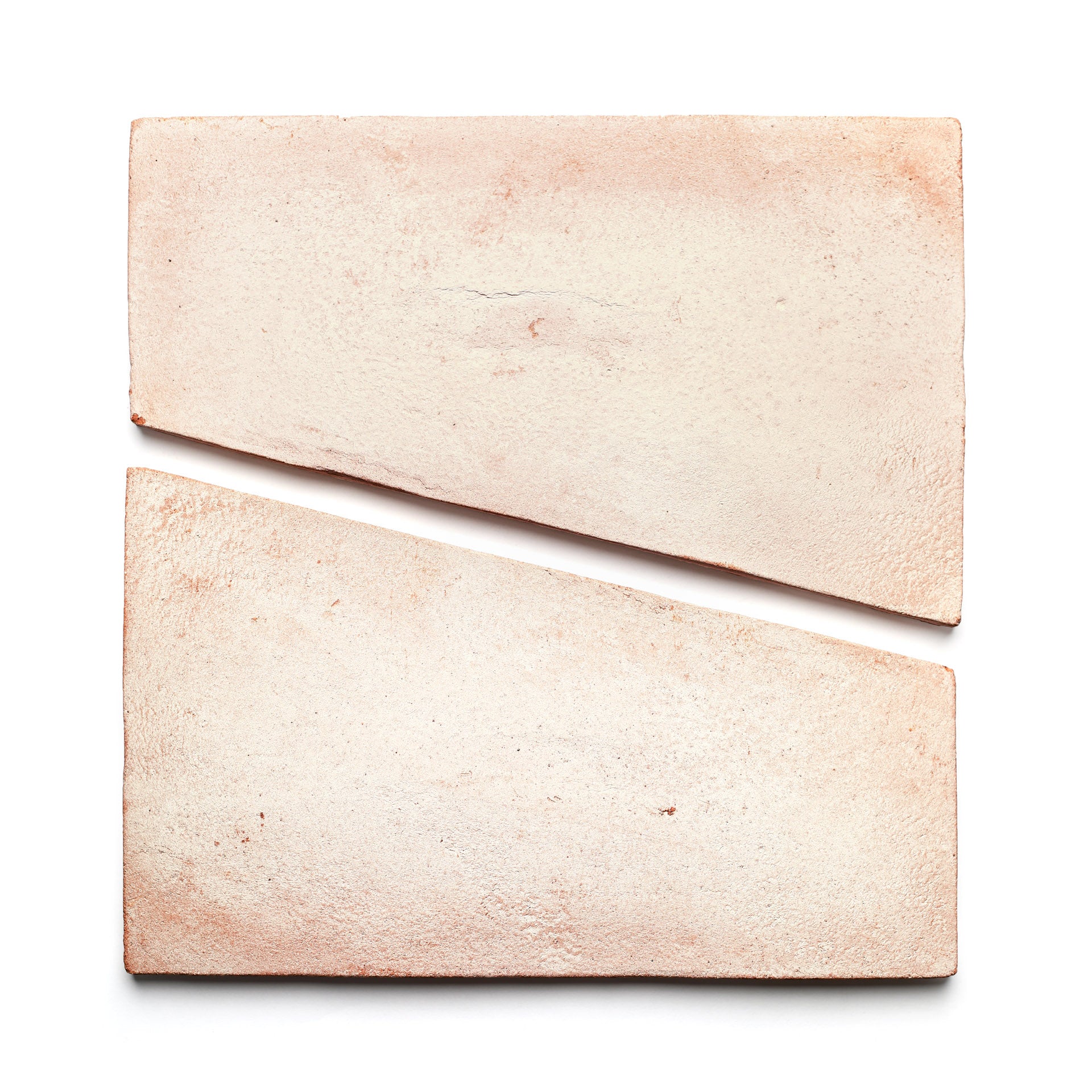 Two Toltec + Blanco tiles by Zia Tile, featuring a light pink hue and handmade texture, are placed on a white background—one whole and one diagonally cut—to create a striking geometric composition.