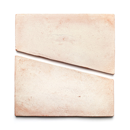 Two Toltec + Blanco tiles by Zia Tile, featuring a light pink hue and handmade texture, are placed on a white background—one whole and one diagonally cut—to create a striking geometric composition.
