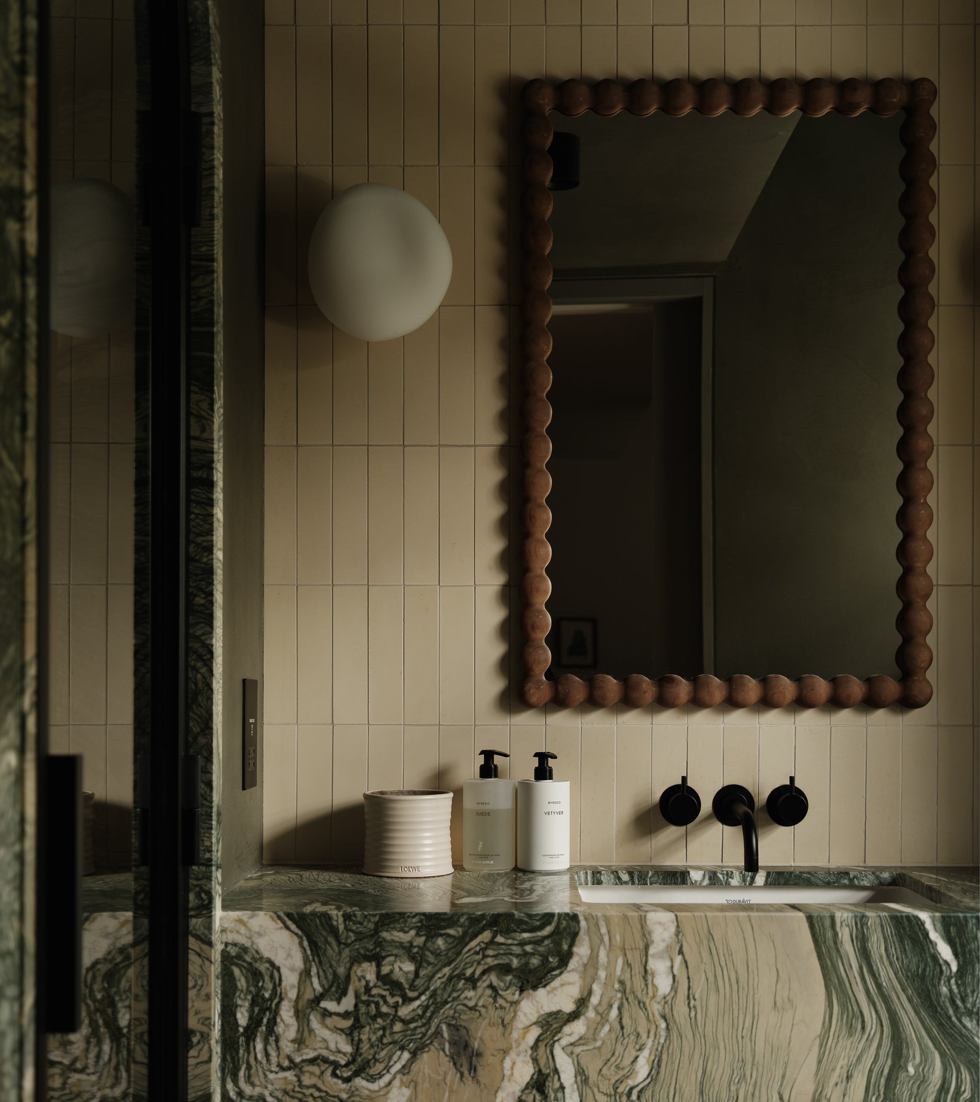 A modern bathroom features beige tiled walls, a large wood-framed mirror, green marble countertop and sink, two soap dispensers, a white cup, a round wall light, matte black fixtures, and Bone 2x8 tile by vendor-unknown.