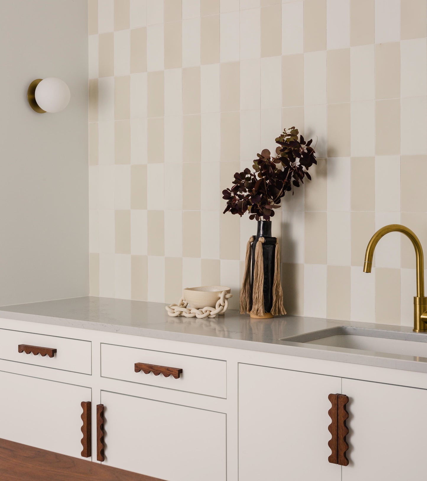 Minimalist kitchen with cream and white checkered Bone 4x8 backsplash tile (vendor-unknown), white cabinets with wavy wood handles, a gold faucet, and décor including dark brown leaves in a vase and an off-white ceramic chain.