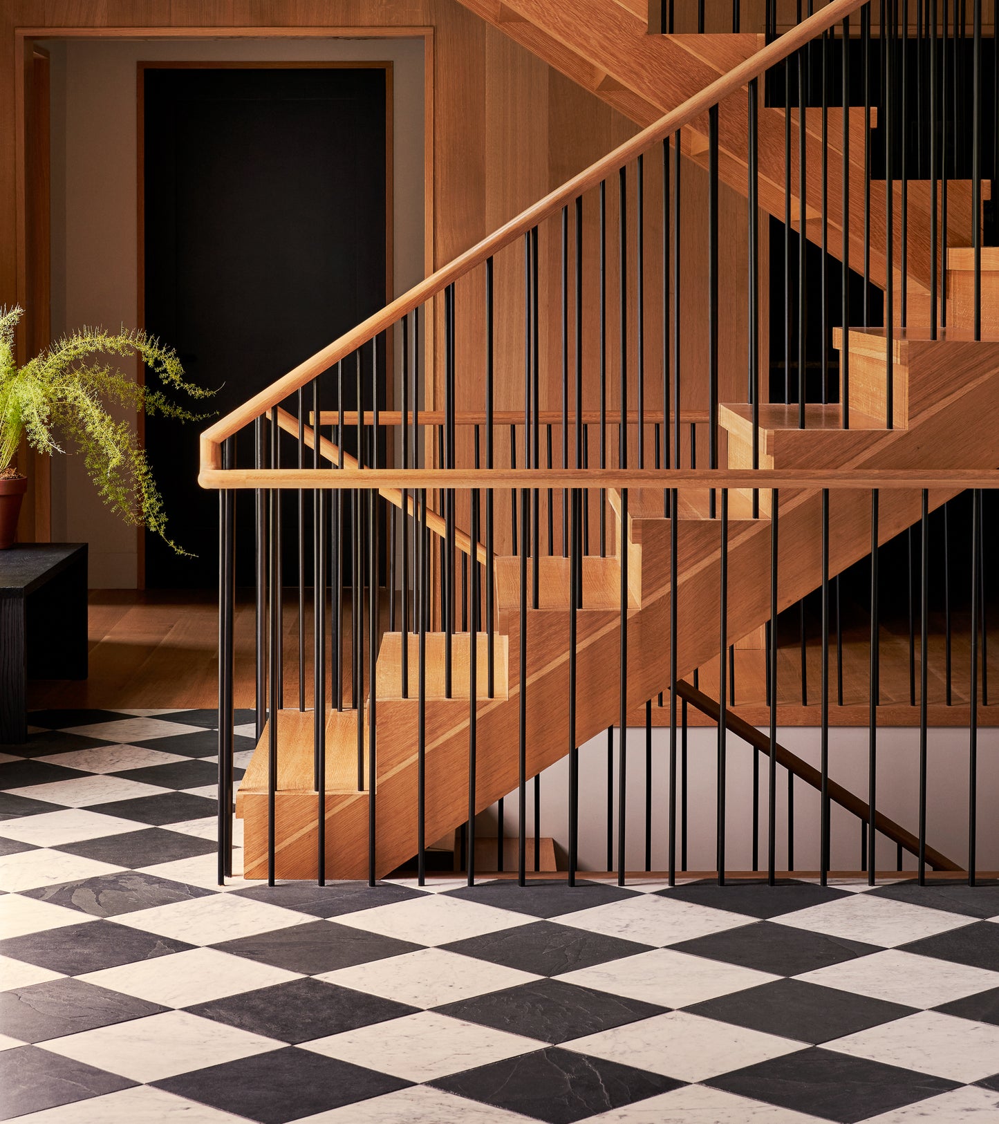 A modern wooden staircase with black railings rises above a Carrara 12x12 floor by Zia Tile; a potted fern sits on a bench to the left, and a black door is visible in the background.