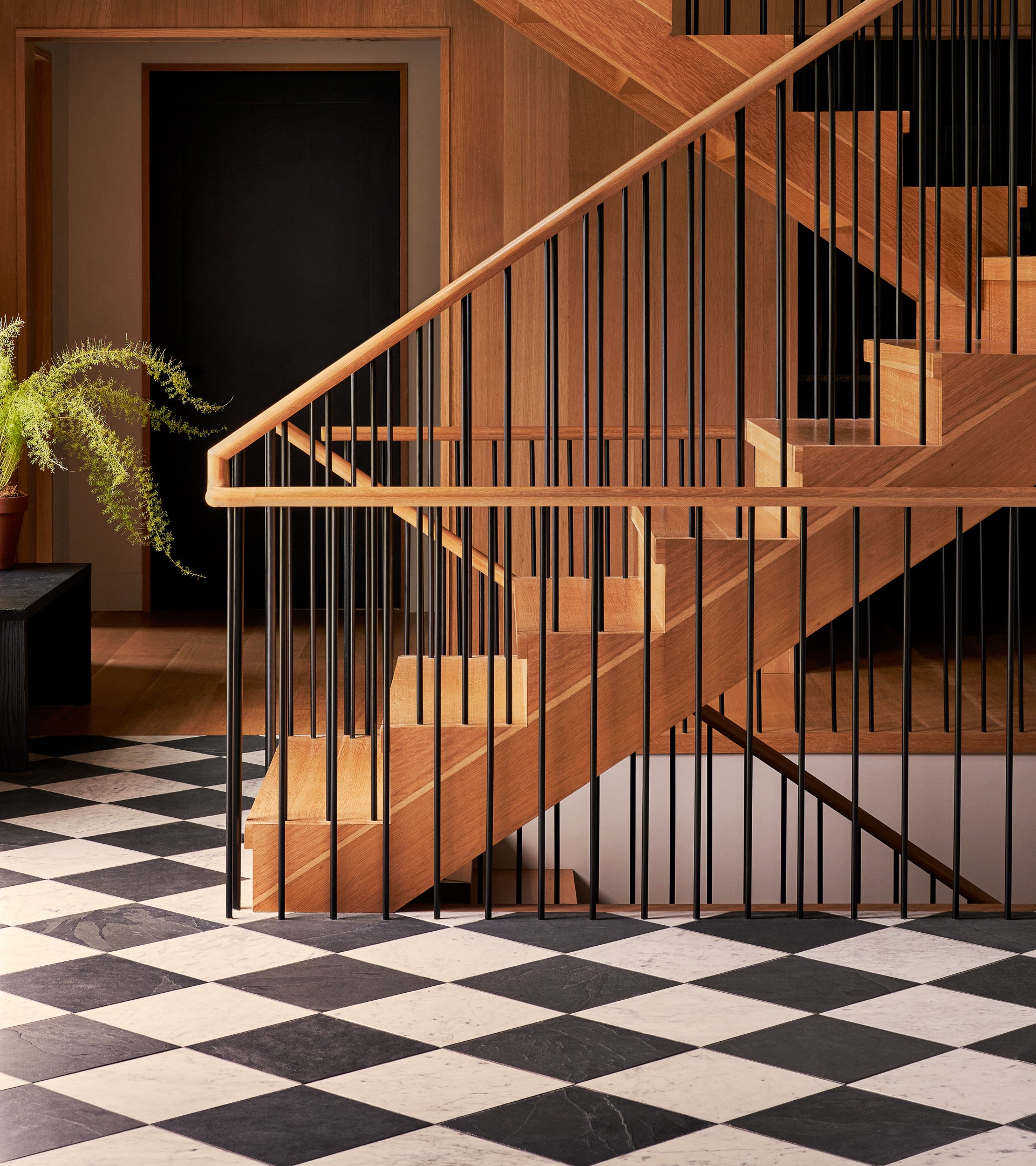 A modern wooden staircase with black railings rises above a Carrara 12x12 floor by Zia Tile; a potted fern sits on a bench to the left, and a black door is visible in the background.