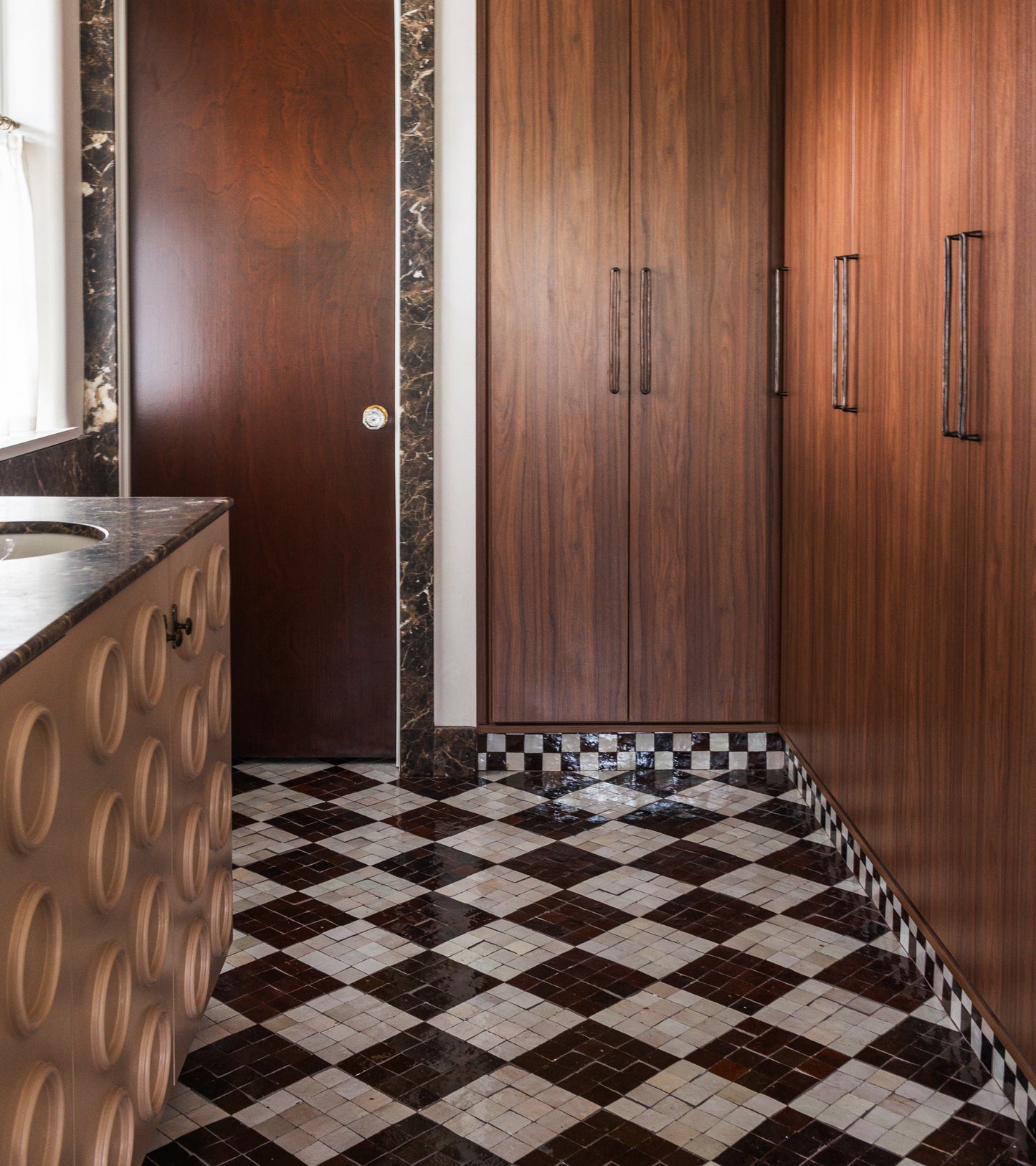 A bathroom with wood-paneled cabinets, a dark wood door, a marble countertop with circular patterns, and Zia Tile's Casablanca 2x2 tiles in black, white, and brown geometric designs. Marble trim accents the door and cabinetry as natural light enters.