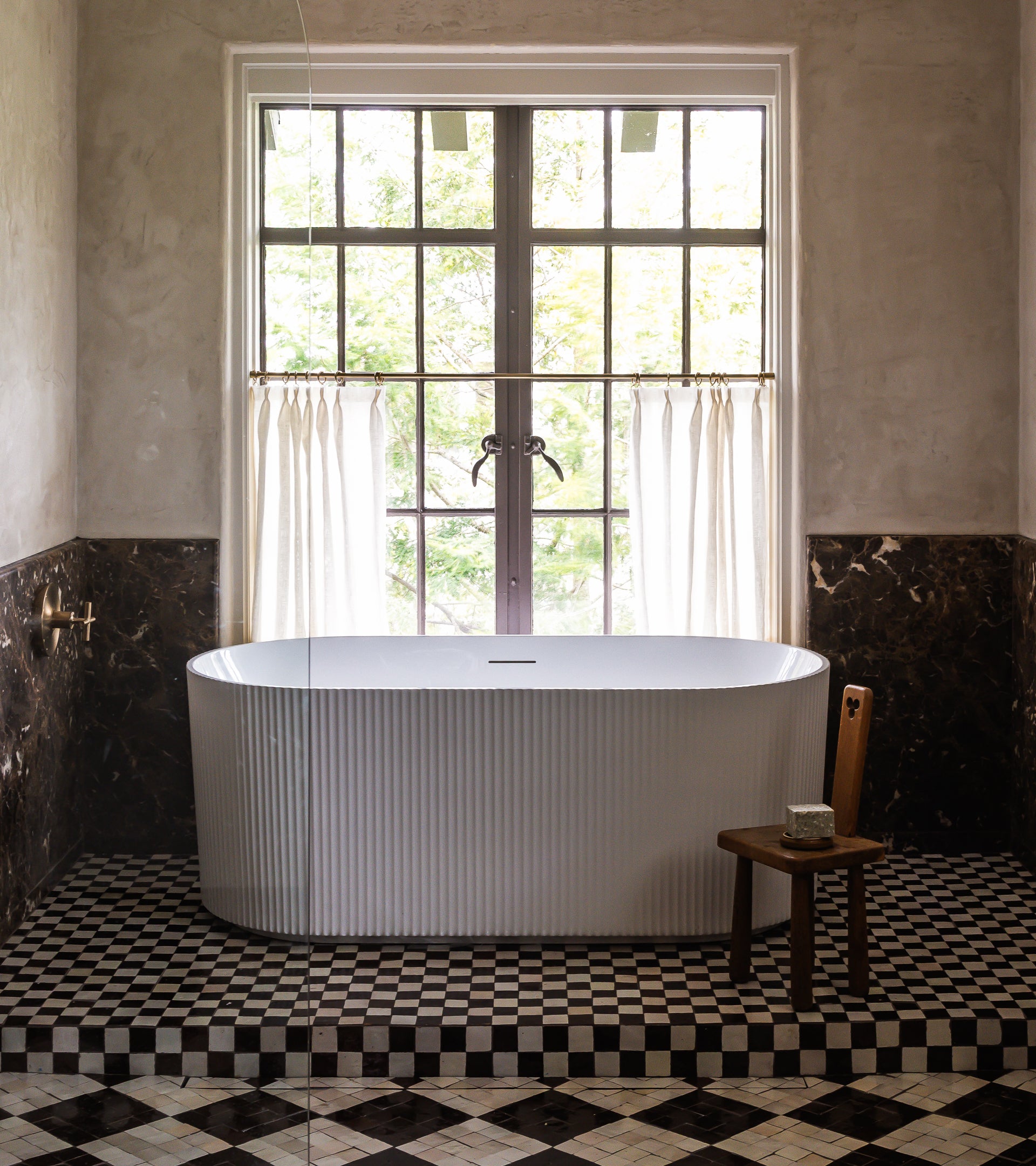 A freestanding white bathtub sits by a large window with sheer curtains, featuring Zia Tile's Casablanca 2x2 Moroccan tile floor, dark marble lower walls, and a wooden chair holding a candle beside the tub.