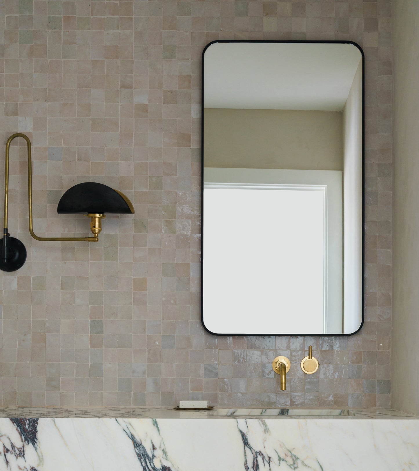 A modern bathroom features a rectangular black-framed mirror, gold wall-mounted faucet, marble countertop, and a brass wall sconce with a half-dome black shade set against Zia Tile's Casablanca 2x2 tile wall.