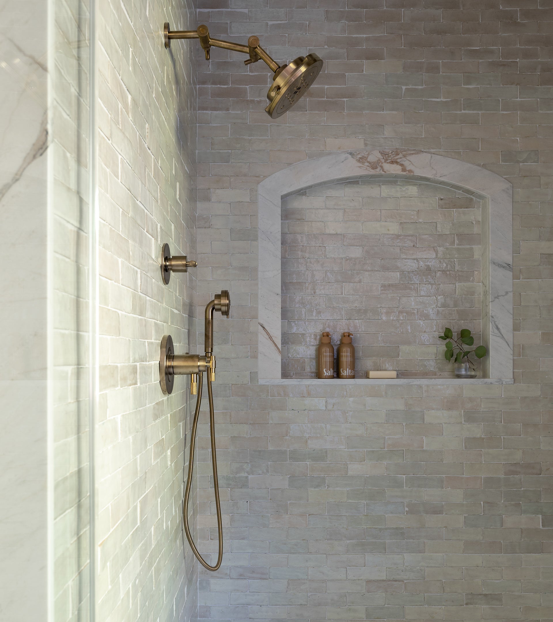 A modern shower features brass fixtures, a handheld and large round showerhead. The wall is lined with Zia Tiles Casablanca 2x6 tiles, while a built-in niche holds two brown bottles, soap, and a small green plant.
