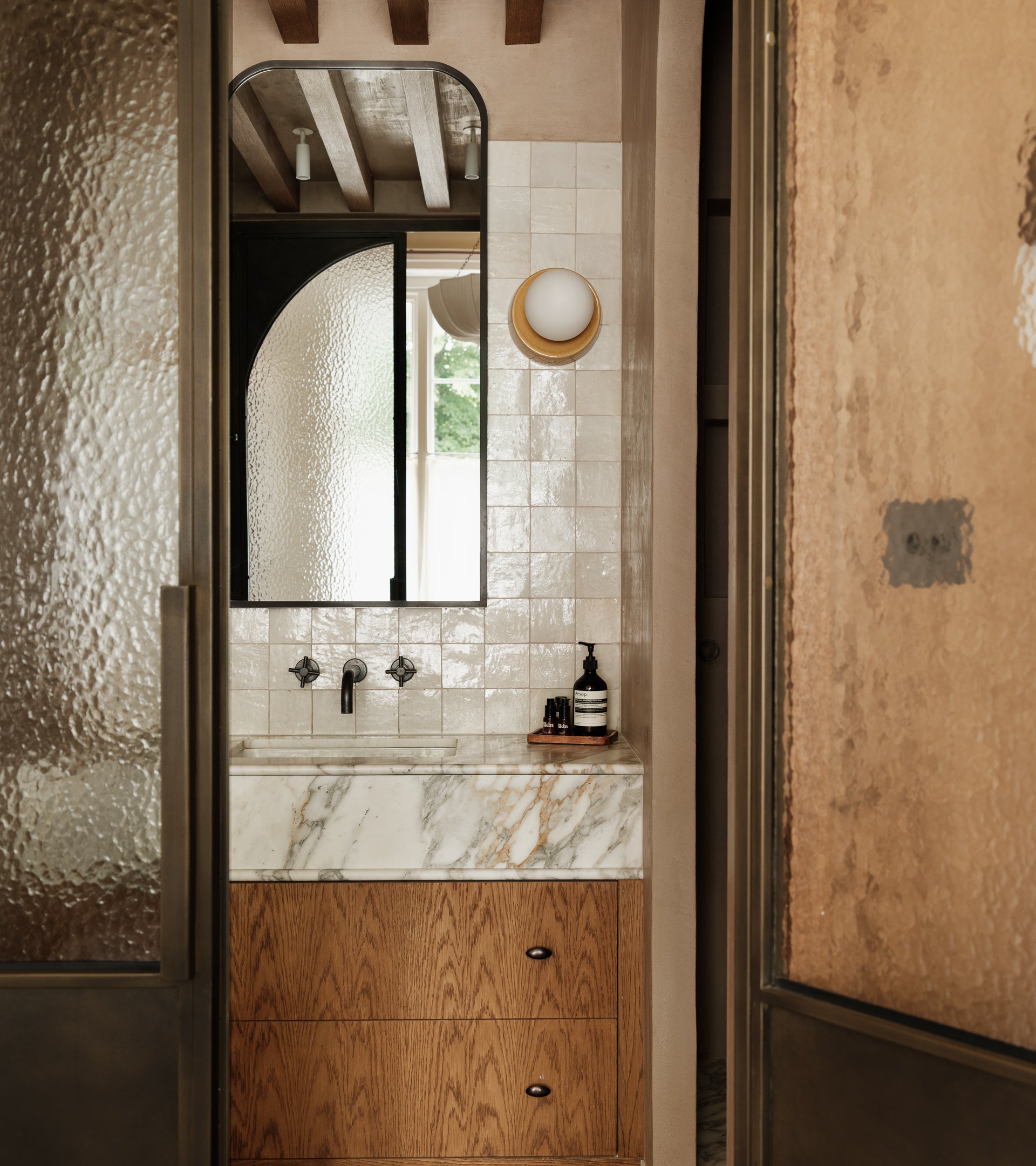 A modern bathroom features a marble sink, wooden cabinetry, and white tiled walls. Frosted glass doors and a round sconce pair beautifully with vendor-unknown Casablanca 4x4 tile for a minimalist, elegant look.