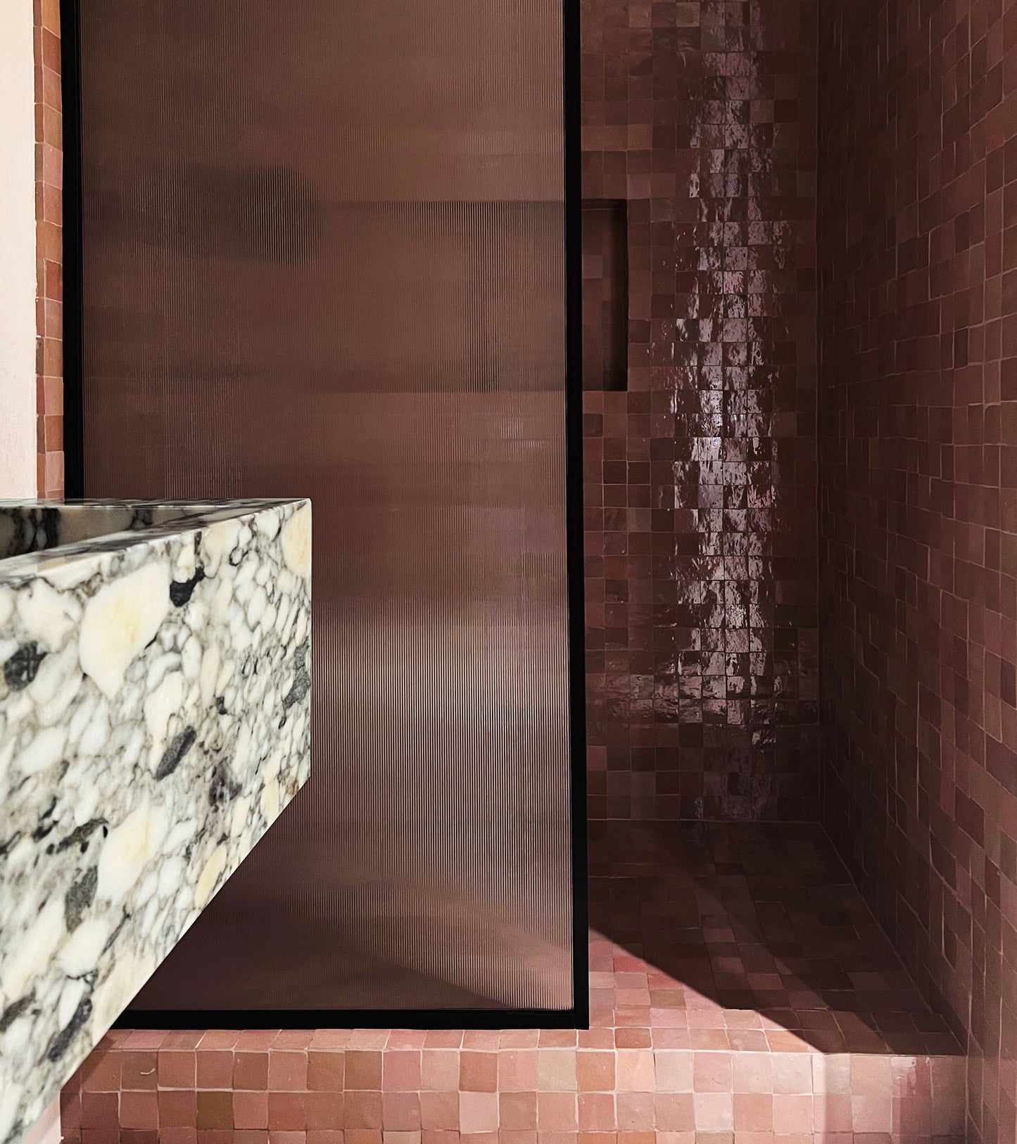 A modern bathroom features Zia Tile's Desert Bloom 2x2 tiles, a marble sink, and a glass shower divider. Light bounces off the glossy Moroccan tiles, filling the space with warmth and style.
