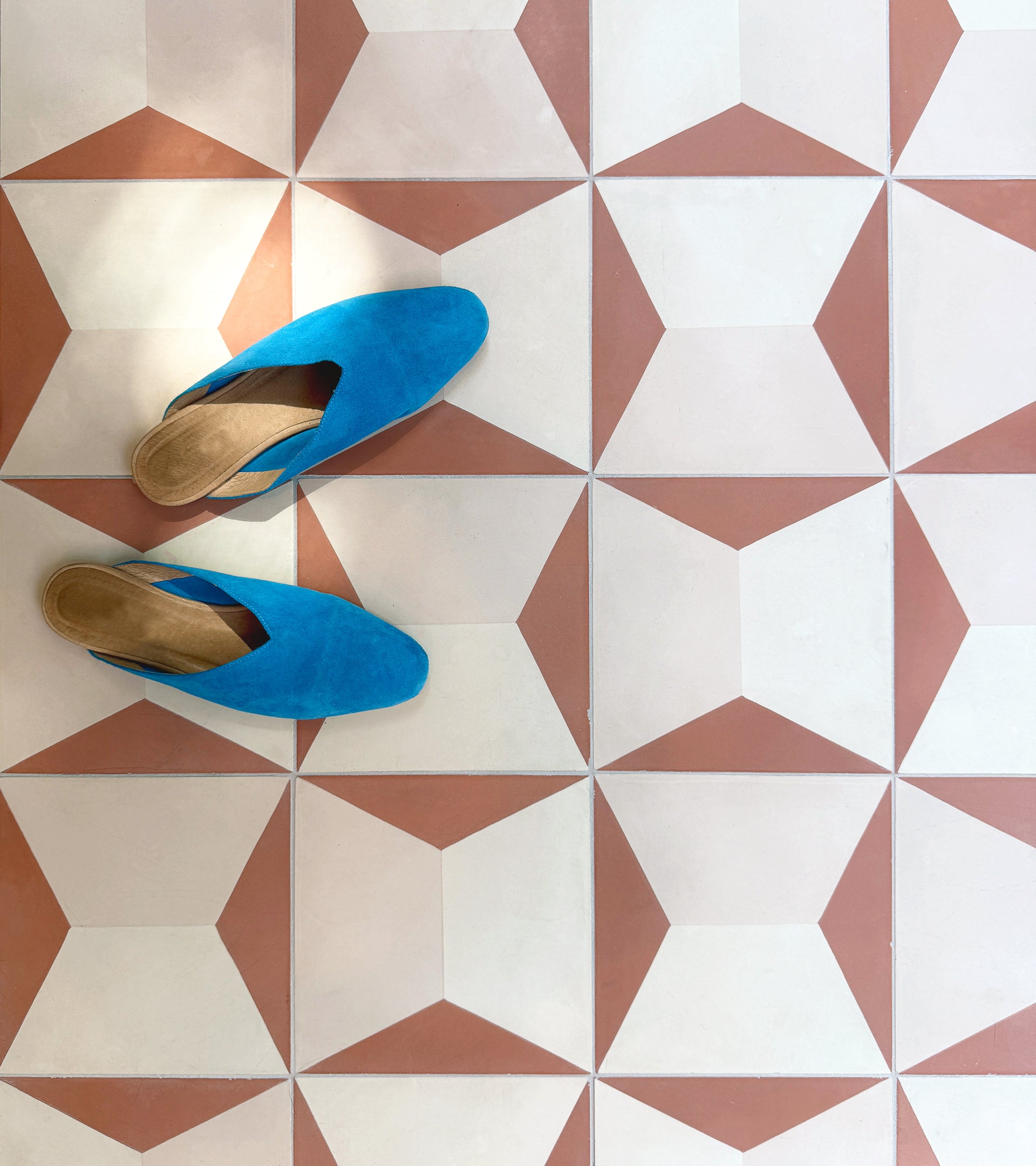 A pair of bright blue slip-on shoes rest on a floor tiled with Zia Tile’s Colorama Dune 8x8, showcasing a geometric pattern in white and terracotta cement tiles.