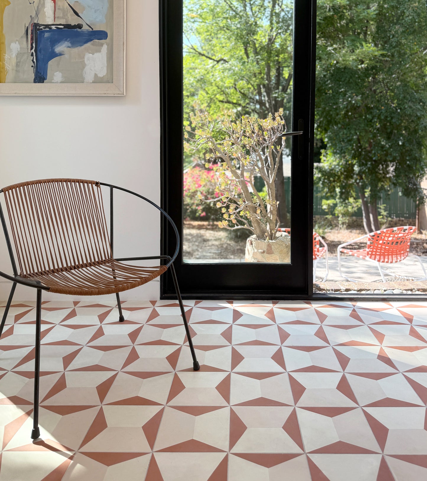 A modern chair with a curved metal frame sits on Zia Tile’s Colorama Dune 8x8 handmade tiles near a glass door, sunlight streaming in and revealing a view of trees and red outdoor chairs outside.
