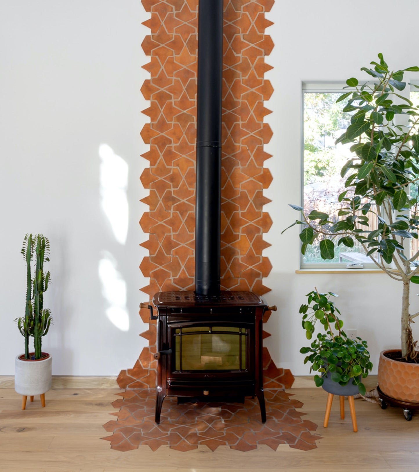 A wood-burning stove with a black chimney sits against a white wall, surrounded by Alcazar + Fired Earth star-shaped terracotta tiles from Zia Tile. Three potted plants and a window bring greenery and natural light to the space.