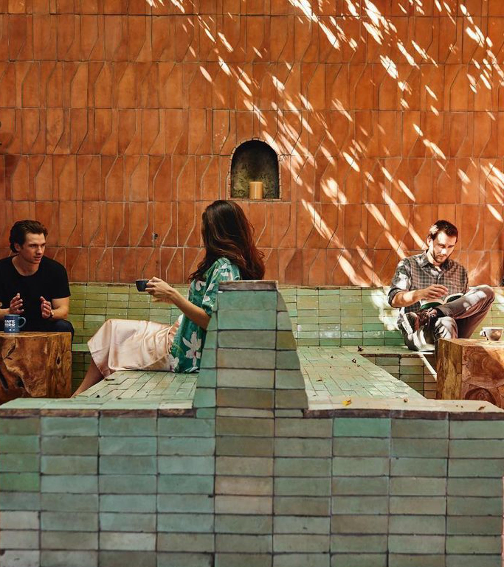 Three people relax on Zia Tile’s Zocalo + Fired Earth brick benches and walls in a sunlit patio. One reads, another sips from a cup, and the third gestures while talking as dappled light plays across the surface.