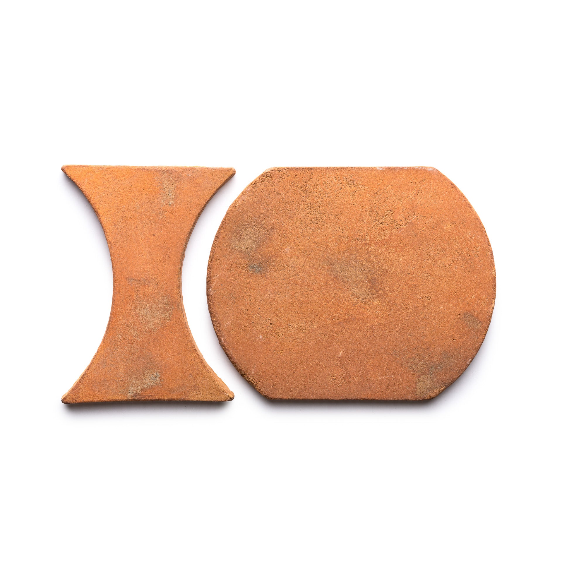 Two reddish-brown terracotta tiles from Zia Tile’s Oaxaca + Fired Earth collection rest on a white background—one hourglass-shaped, the other a rounded rectangle.