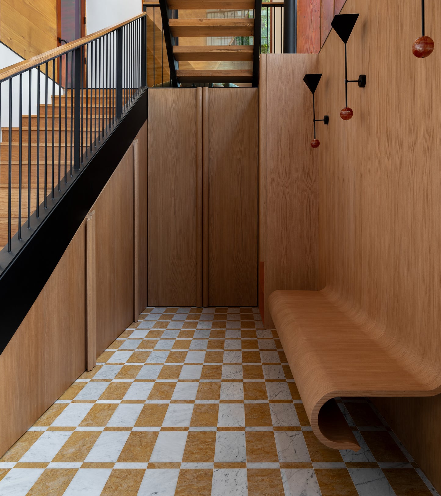 A modern hallway with wood-paneled walls, a curved bench, Zia Tile’s Verona in Carrara + Giallo Reale handcrafted checkered tile floor, a black metal staircase, and black wall lights featuring red glass accents.