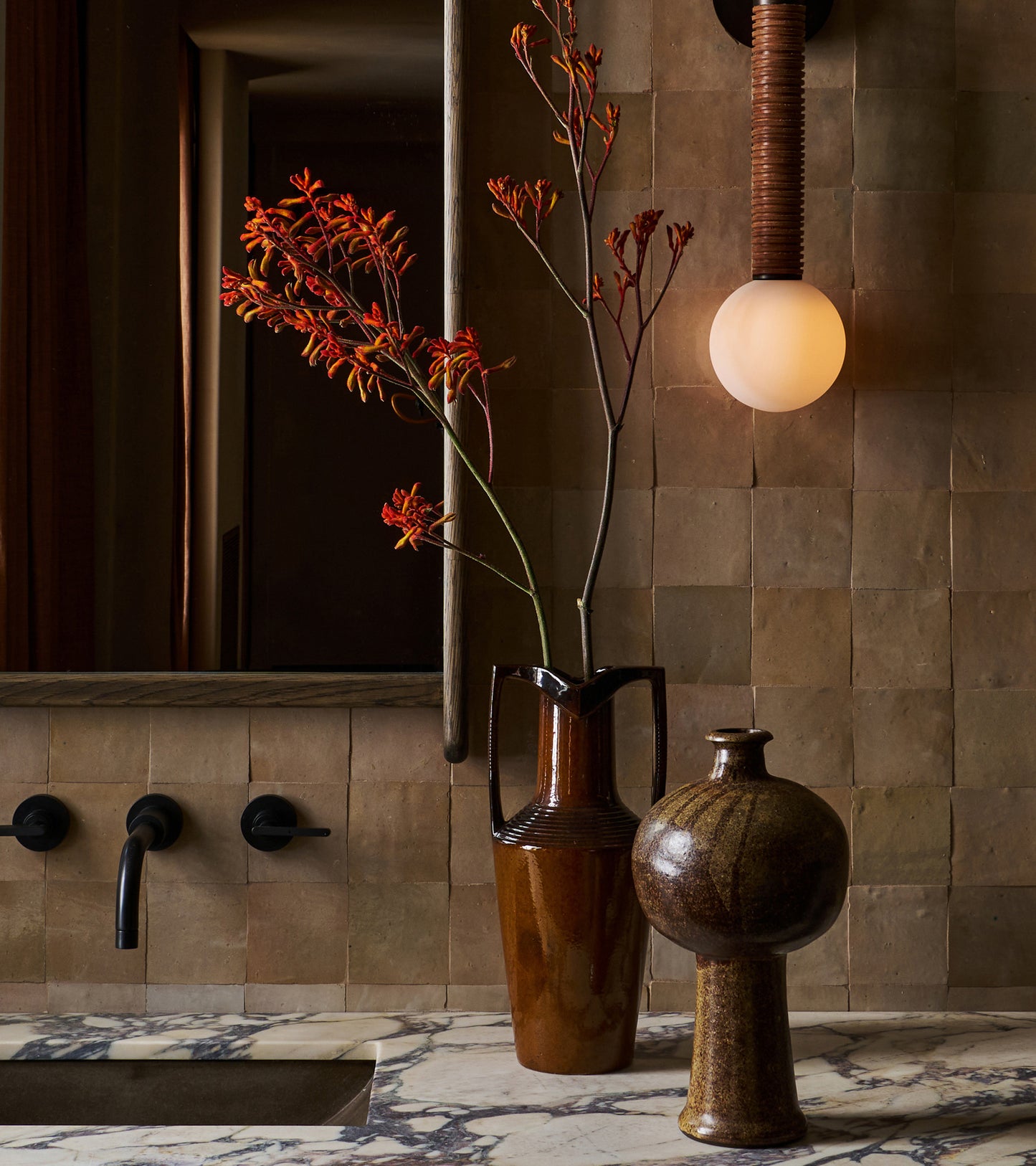 A marble bathroom countertop with a tall brown vase of red-orange flowers, a round ceramic vessel, wall-mounted faucet, and glowing round light sits against vendor-unknown’s Glazed Earth 4x4 tiles.