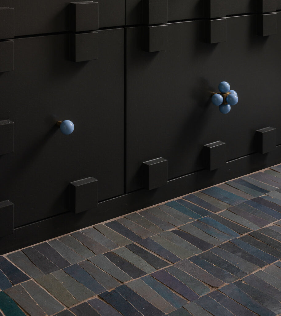 Close-up of black cabinets with square black handles and two round blue knobs, set against a floor of Graphite Grey Trapezoid tiles and green tiles in a staggered pattern for a subtle Moroccan vibe.