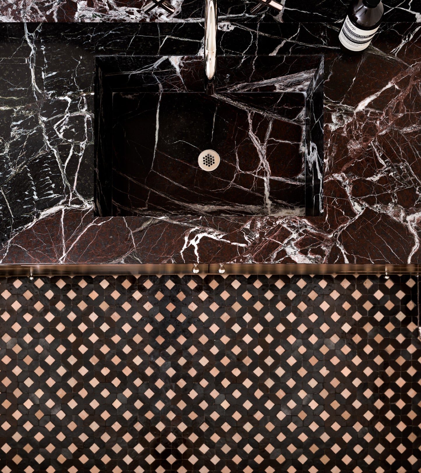 A black marble sink with white veining sits below a silver faucet, atop a matching countertop. The bathroom backsplash features Zia Tile's Lattice 6—a geometric pattern in black, brown, and beige Moroccan tile.