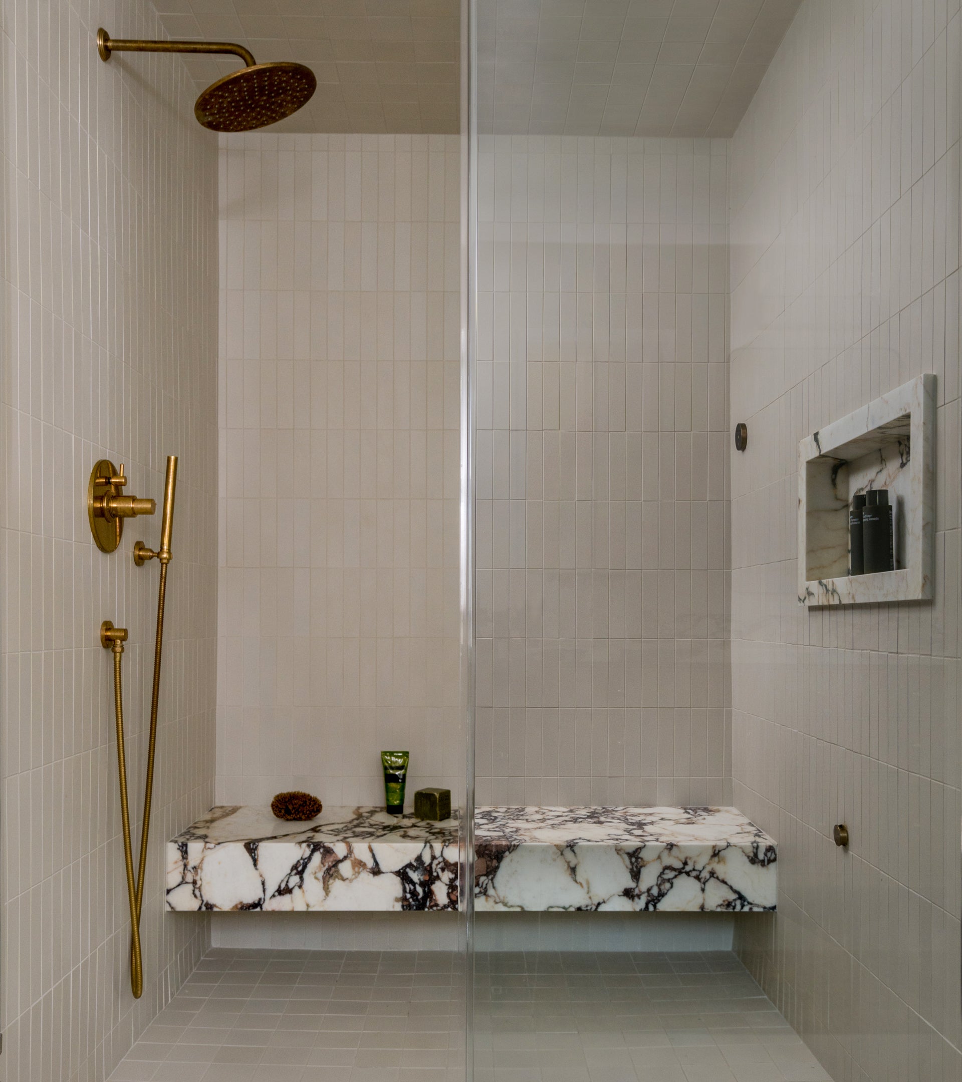 A modern shower features Linen 2x8 ceramic tile walls by Zia Tile, a gold rainfall showerhead and controls, a marble bench, and a built-in niche for toiletries, all presented in a minimalistic and refined design.