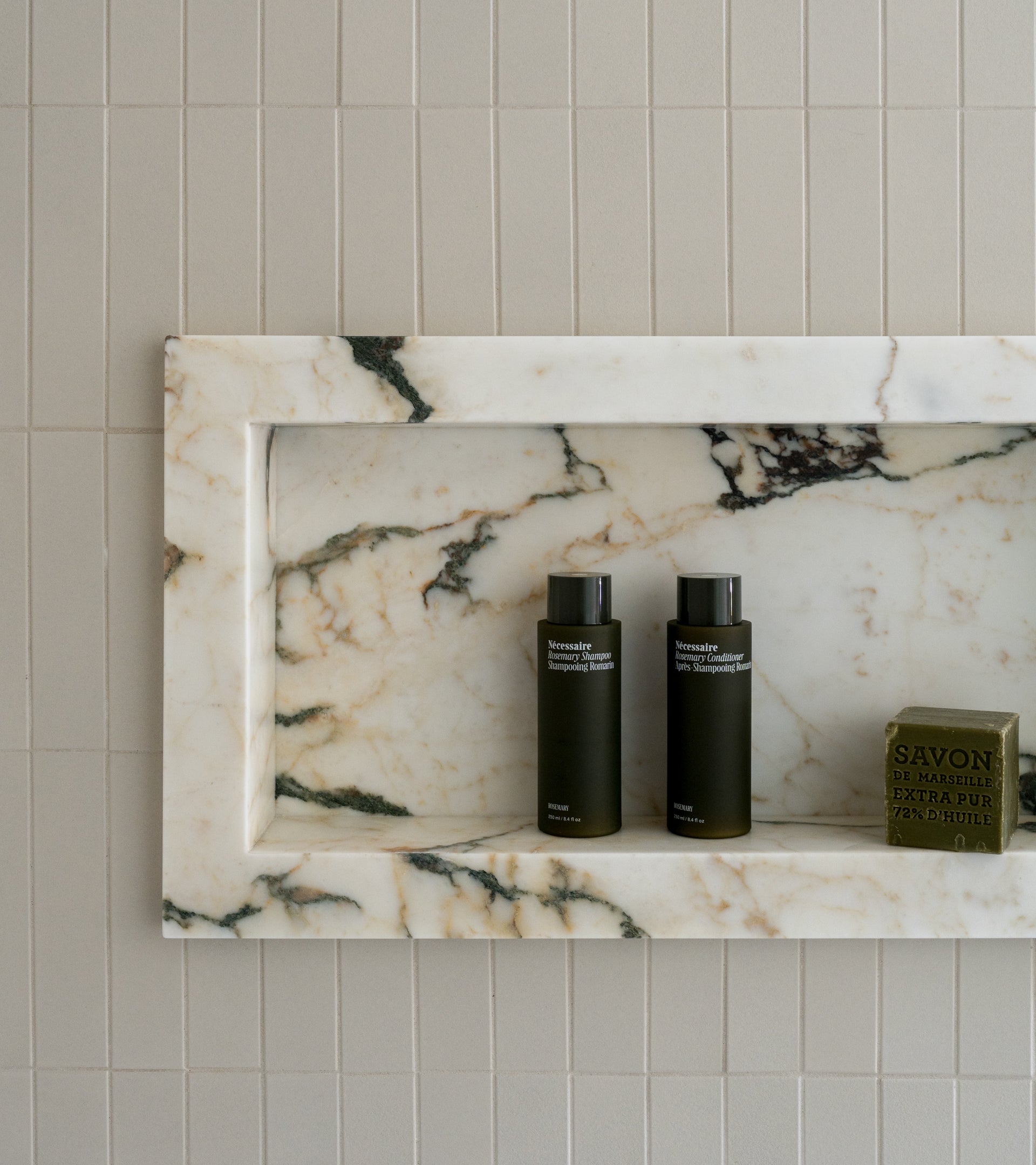 A marble shower niche with two dark bottles and a green bar of soap is set against an elegant wall of Zia Tile’s Linen 2x8 ceramic tile.